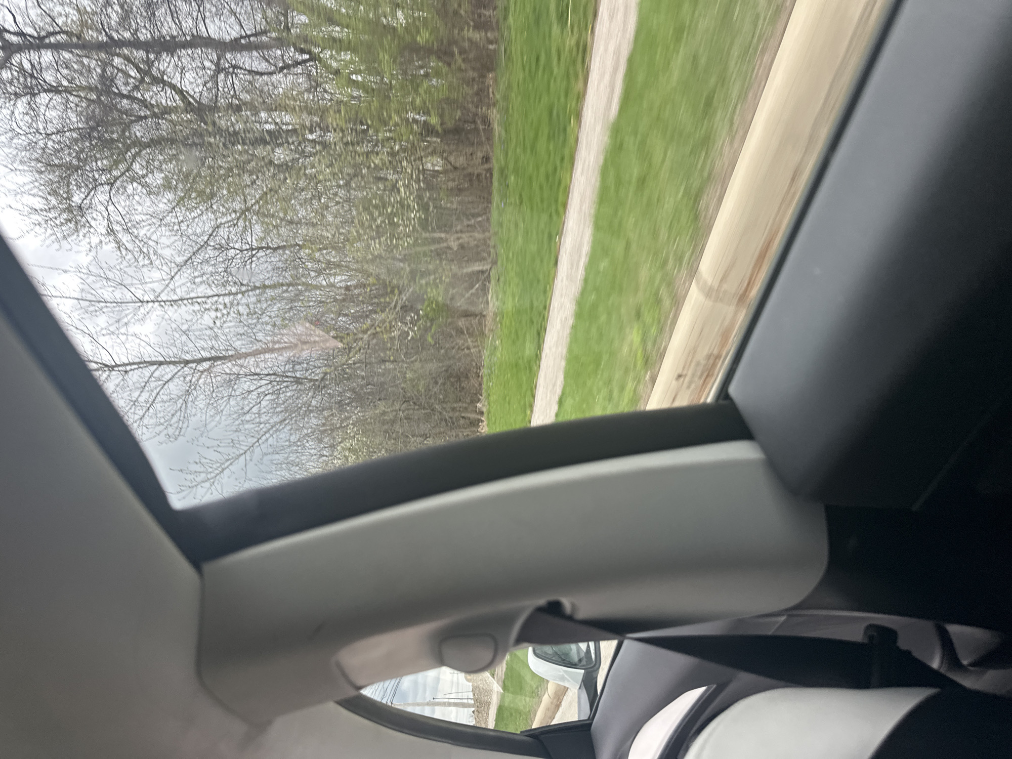 A view from inside a moving car, likely in Pepper Pike, Ohio, USA, shows a blurred landscape through the sunroof and side window. Bare trees line the road, alongside a strip of green grass and a paved path. Interior car elements visible include the headliner, trim, rearview mirror, seat, and seatbelt.
