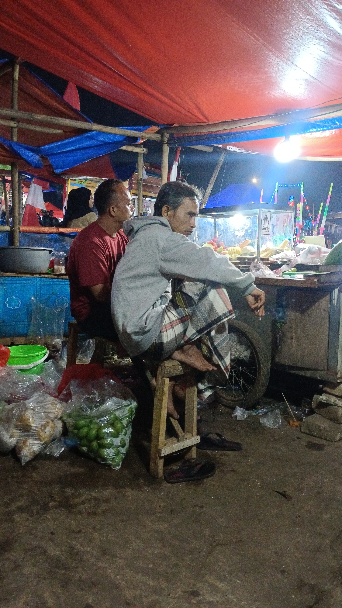 Two men are seated at a food stall under a red and blue tarp structure. The man on the left, wearing a red t-shirt, is looking to the right. The man on the right, wearing a grey hooded sweatshirt and a checkered sarong, is looking forward and appears to be smoking a cigarette. He is seated on a wooden stool. In front of the stall, there are bags of produce, including a bag filled with green fruits. To the right of the stall, a tire is visible. The ground is a dirt surface. The background shows the general environment of a market or roadside stall at night, with lights and other stalls suggested. The scene is located in Tajurhalang, Indonesia.