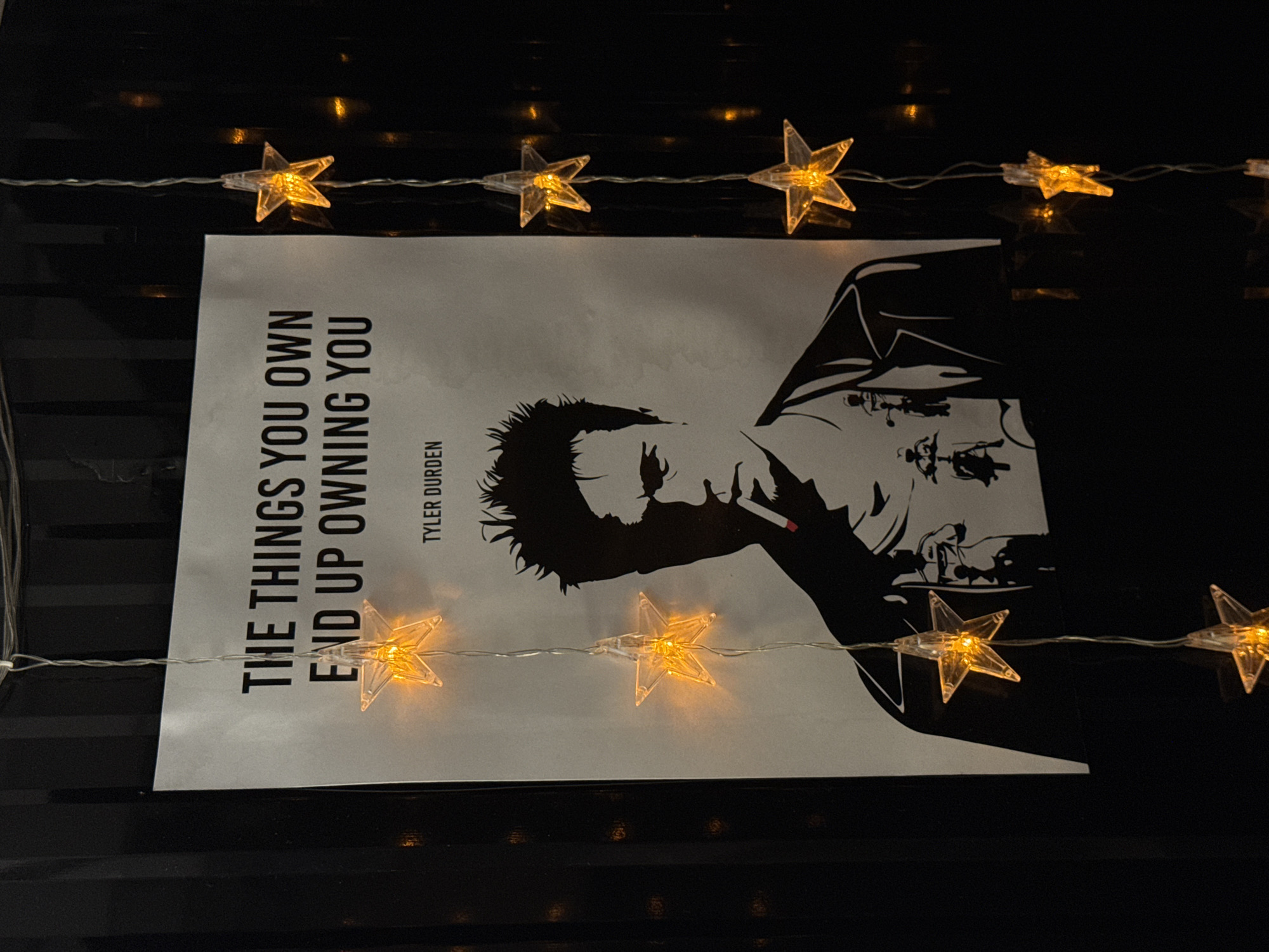 A framed poster of Tyler Durden from the movie Fight Club is displayed on a dark, ribbed wall, possibly a black metal corrugated fence or similar structure. The poster features Durden's silhouette, smoking a cigarette, with the quote "THE THINGS YOU OWN END UP OWNING YOU" printed above his head. String lights with illuminated star-shaped bulbs are draped over the poster, casting a warm orange glow. The lights hang vertically on the right side of the poster and also partially obscure the top left corner. The surrounding environment is dark, with reflections visible on the dark surface behind the poster. The image appears to be taken indoors or in a sheltered outdoor space. No specific location in Phuket City Municipality, Thailand can be identified from the visual information provided.