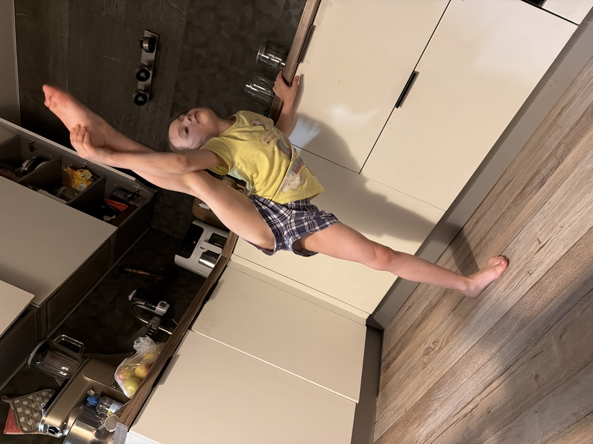 A young girl performs an impressive full split in what appears to be a modern kitchen. The image is rotated 90 degrees clockwise, making her appear to be doing a vertical split between the kitchen elements.
The girl, a child, is dressed in a yellow t-shirt featuring a rainbow graphic and blue and white plaid shorts. She holds her raised left leg with both hands, extending it towards a dark, textured tiled wall, while her right leg stretches downwards towards a light wooden floor. Her head is tilted back slightly, and she looks upwards, demonstrating focus and flexibility.
The setting is a well-lit kitchen, characterized by sleek, light-colored cabinets with minimal black handles. An open lower cabinet/drawer on the left side reveals various kitchen items, including a metallic food mixer, a plastic bag containing several apples, and a water bottle. Above where the girl's left hand is gripping, two glass cups are visible on a shelf or counter. The floor is covered with light-toned wooden planks. No visible text is clearly legible beyond the graphic on the girl's shirt. The scene takes place indoors, in Perm, Russia.