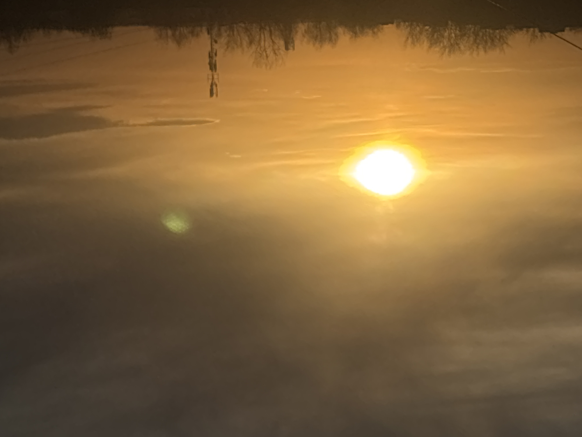 The scene depicts a sunrise or sunset over Chaikovsky, Russia. The sky displays a gradient of colors, transitioning from bright golden-orange in the lower and mid-sections, illuminated by the sun, to a darker grey-brown in the upper-right portion. The sun is prominently visible as an intense, overexposed white orb with a broad golden glow, located in the upper-left quadrant of the frame. A distinct green, hazy oval artifact, consistent with a lens flare, is present in the upper-right sky. Scattered, thin clouds are observed throughout the illuminated areas, with denser cloud formations in the darker upper-right.
The lower portion of the image features a dark, silhouetted horizon line. This silhouette reveals a continuous band of bare deciduous trees, indicative of winter or early spring. Rising above the tree line in the right-center is a tall, slender communication tower, clearly visible in silhouette with multiple antenna arrays attached. Faint, dark linear elements, consistent with utility wires, extend diagonally upwards from the lower-left foreground. No individuals are visible.