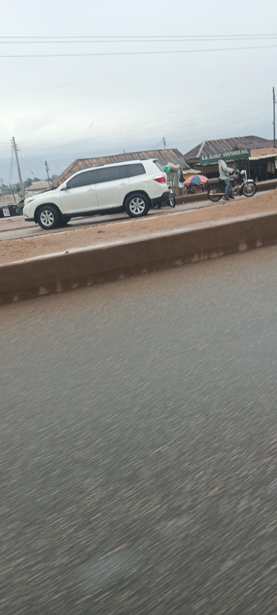 The image captures a roadside scene in Bukuru, Nigeria, from the perspective of a moving vehicle, evidenced by the motion blur on the asphalt road in the foreground. The main subjects are a white Toyota Highlander SUV and a motorcycle with a rider, both positioned on the side of the road.
The white SUV is prominently featured, parked or stationary, facing left in the frame. To its right, a person wearing a light-colored hoodie or jacket sits on a motorcycle, which also appears to be stationary or moving slowly. Further behind the motorcycle, an open umbrella hints at a small vendor stall or shaded area.
In the background, several single-story structures with corrugated iron roofs line the road. On one of these buildings, a sign is clearly visible, displaying the text "B.B. GLOBAL VENTURES NIG." The abbreviation "NIG." likely refers to Nigeria. Utility poles with overhead lines are visible against the sky, indicating an established infrastructure. The overall sky is overcast and gray, suggesting either an early morning, late afternoon, or simply a cloudy day. The setting appears to be a typical commercial or residential area along a main road in a Nigerian town.