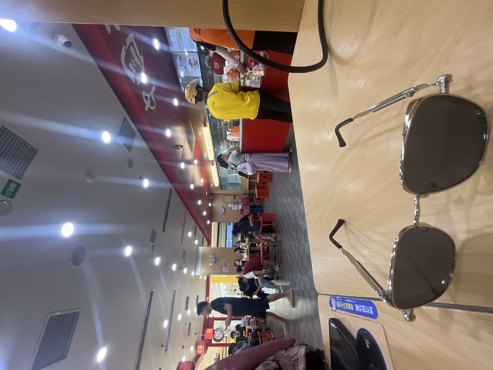 The image depicts the interior of a brightly lit fast-food or casual dining restaurant located in Yuzhong District, China. The scene is observed from a low angle at a light wood-grain table, where a pair of silver-framed sunglasses with dark lenses rests prominently in the foreground. To the left of the sunglasses, a black smartphone is partially visible next to a blue vertical strip of paper with Chinese characters. A coiled black cable extends into the frame from the right.
In the mid-ground, the restaurant floor is tiled with dark gray squares. Multiple patrons are present. On the left, a person in a dark red garment is partially visible. Further back, a man in a black T-shirt and black shorts, wearing light-colored slippers, walks from left to right while looking at a smartphone. Behind him, a group of approximately five individuals is seated around a larger table, with red plastic stools, engaged in dining or conversation. One individual in a light blue top and gray pants leans forward towards the table.
In the background, a service counter extends across the width of the frame. A woman in a light-colored top and a long purple skirt, carrying a light-colored backpack, stands at the counter, facing menu displays adorned with Chinese text and food images, one sign prominently featuring "免费送" (Free Delivery) and "100元" (100 yuan). To her right, a delivery driver in a bright yellow T-shirt and a yellow motorcycle helmet stands with his back to the viewer, appearing to wait for an order; a "美团" (Meituan) logo is visible on his shirt. Another individual in an orange T-shirt is partially visible on the far right, reaching over the counter.
The restaurant ceiling is white with numerous recessed circular light fixtures, casting visible light rays. Rectangular air vents are integrated into the ceiling, alongside a circular speaker and a smoke detector. A white CCTV camera is mounted near the red upper wall section above the counter. An upside-down green sign with Chinese characters "新风口" (New Air Outlet) is visible on the ceiling in the upper left. The upper wall section above the counter area is red, featuring lighter-colored decorative elements or logos.