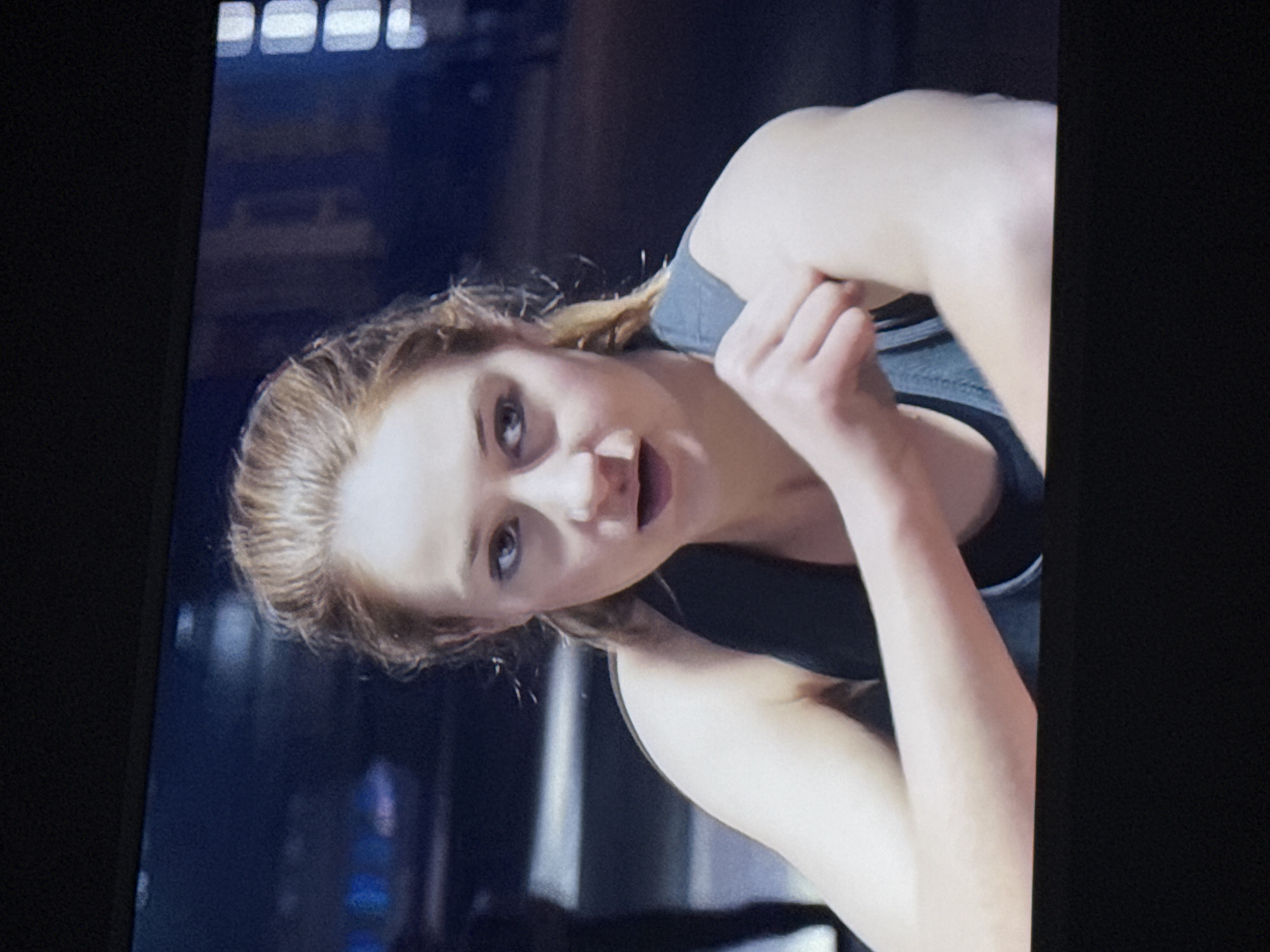 A woman with fair skin, light brown hair pulled back, wearing a black and gray athletic tank top, is seen in a medium close-up shot. Her mouth is slightly open, and she is looking directly at the camera. Her right hand is near her neck. The background is blurry but appears to be an interior space, possibly a gym or fitness studio. The location is not determinable as Mytishchi, Russia.