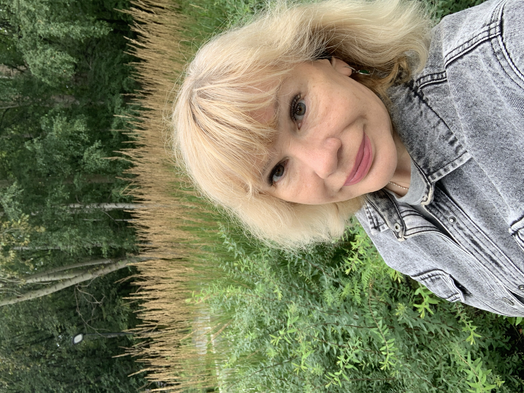 A medium shot captures a blonde woman in a denim jacket, posing outdoors. She is positioned slightly off-center to the right, with her face turned towards the camera. The background consists of dense green foliage in the foreground, transitioning to tall, dry grasses, and finally a line of trees in the distance. The overall lighting suggests an overcast day. The specific location in Prokopyevsk, Russia, is not discernible from the visual information provided in the image.