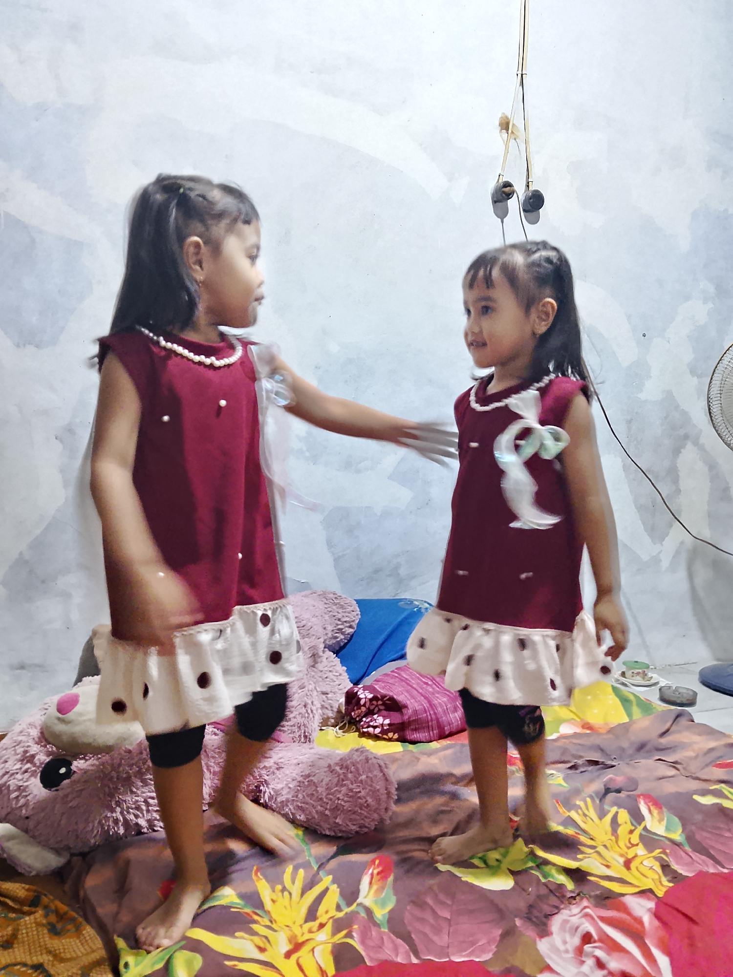 The image depicts two young girls in matching maroon dresses with white, polka-dotted skirts and black leggings, standing on a bed covered with a floral patterned comforter. The girl on the left is facing away from the camera, while the girl on the right is facing towards the camera. Both girls have dark hair styled in ponytails. A large, light purple stuffed bear is visible behind the girl on the left. In the background, a plain, textured wall is visible, with a pair of earphones hanging from a wire suspended from the ceiling. To the right of the girls, a small portion of a fan can be seen. The lighting appears to be artificial, casting shadows on the wall. The scene is set in an indoor environment, possibly a bedroom, in the city of Tanggungan, Indonesia.