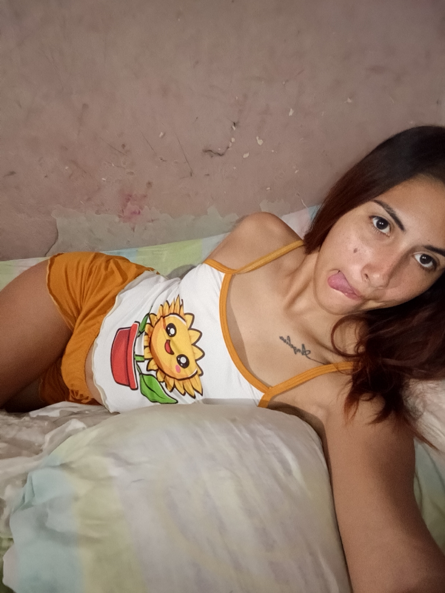 A young woman with dark hair lies on a bed, propped up on her side. She is wearing an orange and white tank top with a sunflower graphic and orange shorts. Her tongue is sticking out as she looks at the camera. There is a tattoo on her chest. The background consists of a light-colored wall and a white pillow. The overall impression is of a casual, indoor setting.