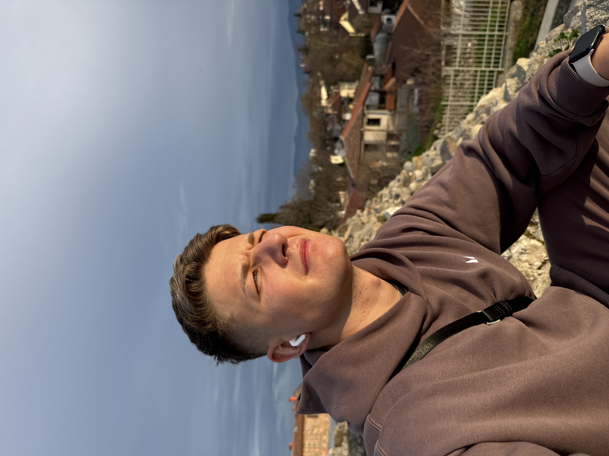 A man is angled in the frame, facing upwards towards the sun, squinting slightly. He is wearing a brown hoodie with a white logo on the chest and has a black strap across his torso. He has short brown hair and is wearing a white wireless earbud in his left ear. His left wrist has a smartwatch. The background shows a cityscape with red-tiled roofs of buildings and a mountain range in the distance under a bright sky. The man is standing on a stone wall. The location is the ancient theatre in Plovdiv, Bulgaria.