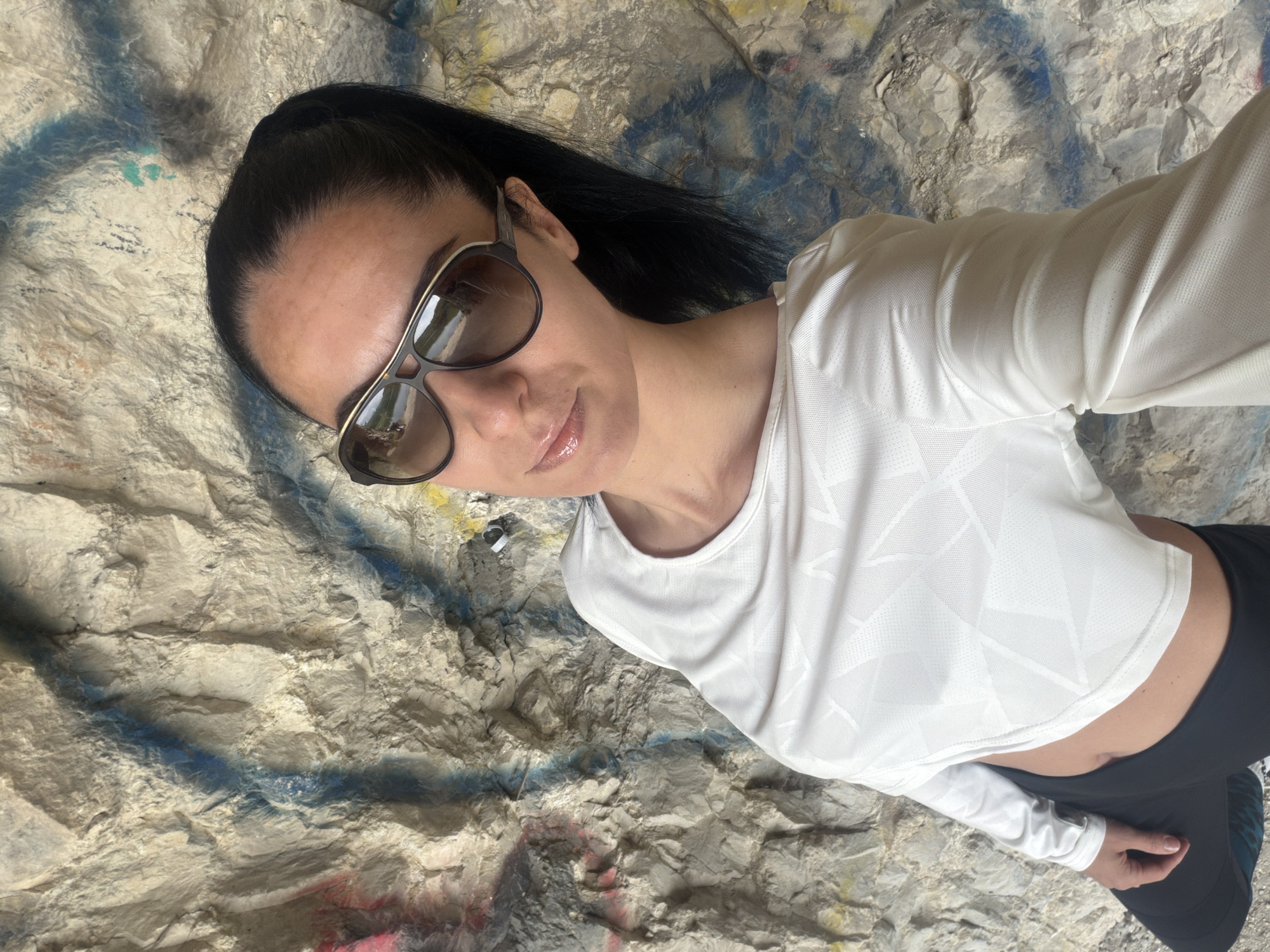 A light-skinned woman with dark hair pulled back in a ponytail, wearing sunglasses, a long-sleeved white shirt with a geometric pattern, and black leggings, is taking a selfie. She is standing in front of a weathered, light-colored rock face with traces of blue graffiti. This location is likely in or near Pleven, Bulgaria, as specified in the prompt.