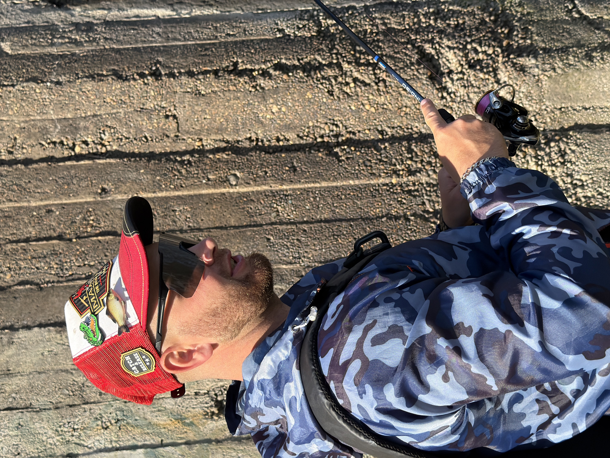 A man is fishing. He is wearing a camouflage jacket, a red and white trucker hat with a fish pin, and sunglasses. He is holding a fishing rod and reel. The background is a concrete structure. This is in the city of Chaikovsky, Russia.