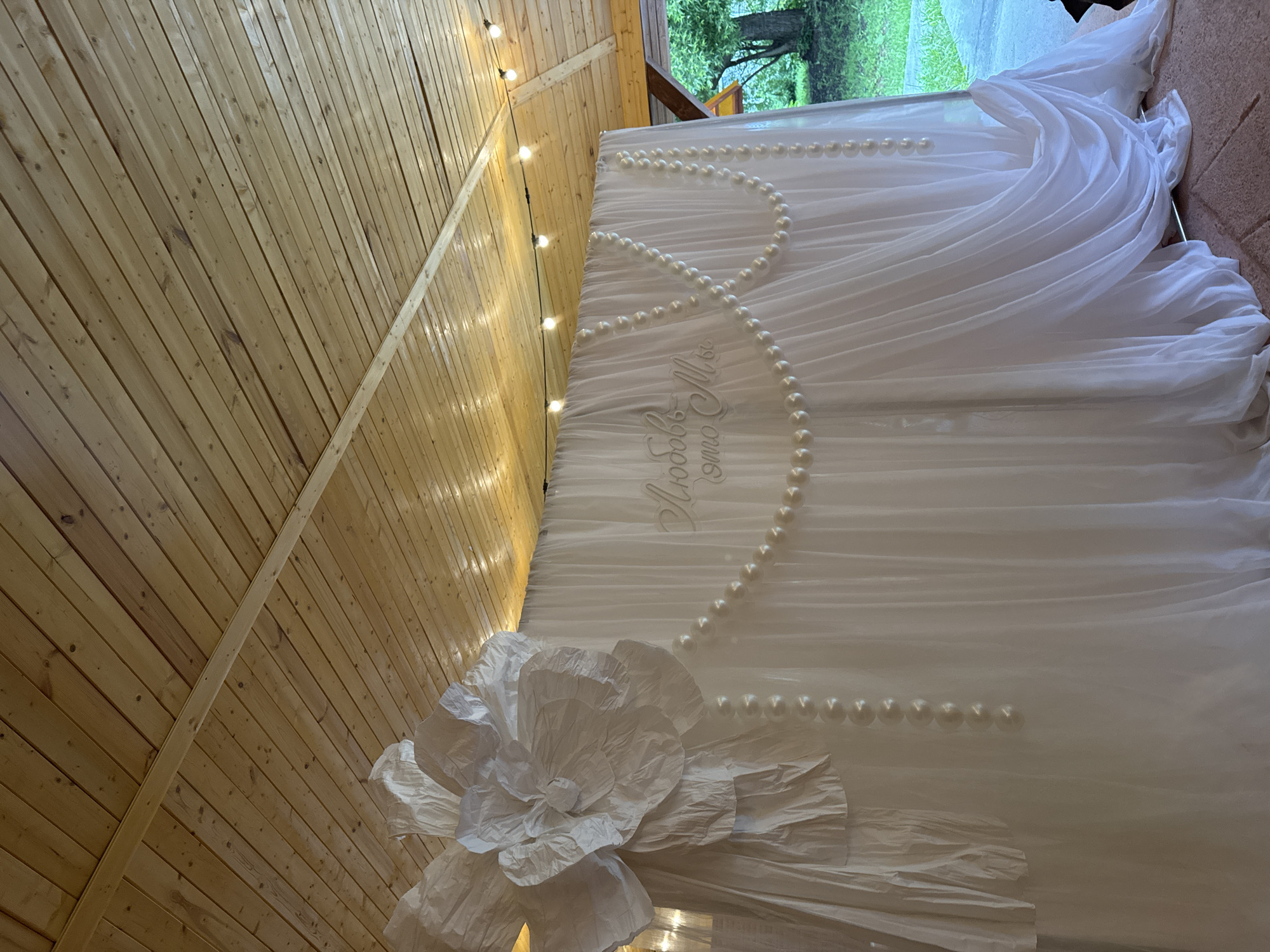 An outdoor wedding decoration scene is depicted. A white sheer fabric backdrop is adorned with a large white paper flower on the left side and draped pearl strands across the center, which feature Cyrillic text that translates to "Love is us." String lights are suspended from a wooden plank ceiling above. The backdrop is set up in an area with a stone-like floor and a paved pathway, with lush green trees visible in the background. The photograph appears to have been taken in Vereshchagino, Russia.