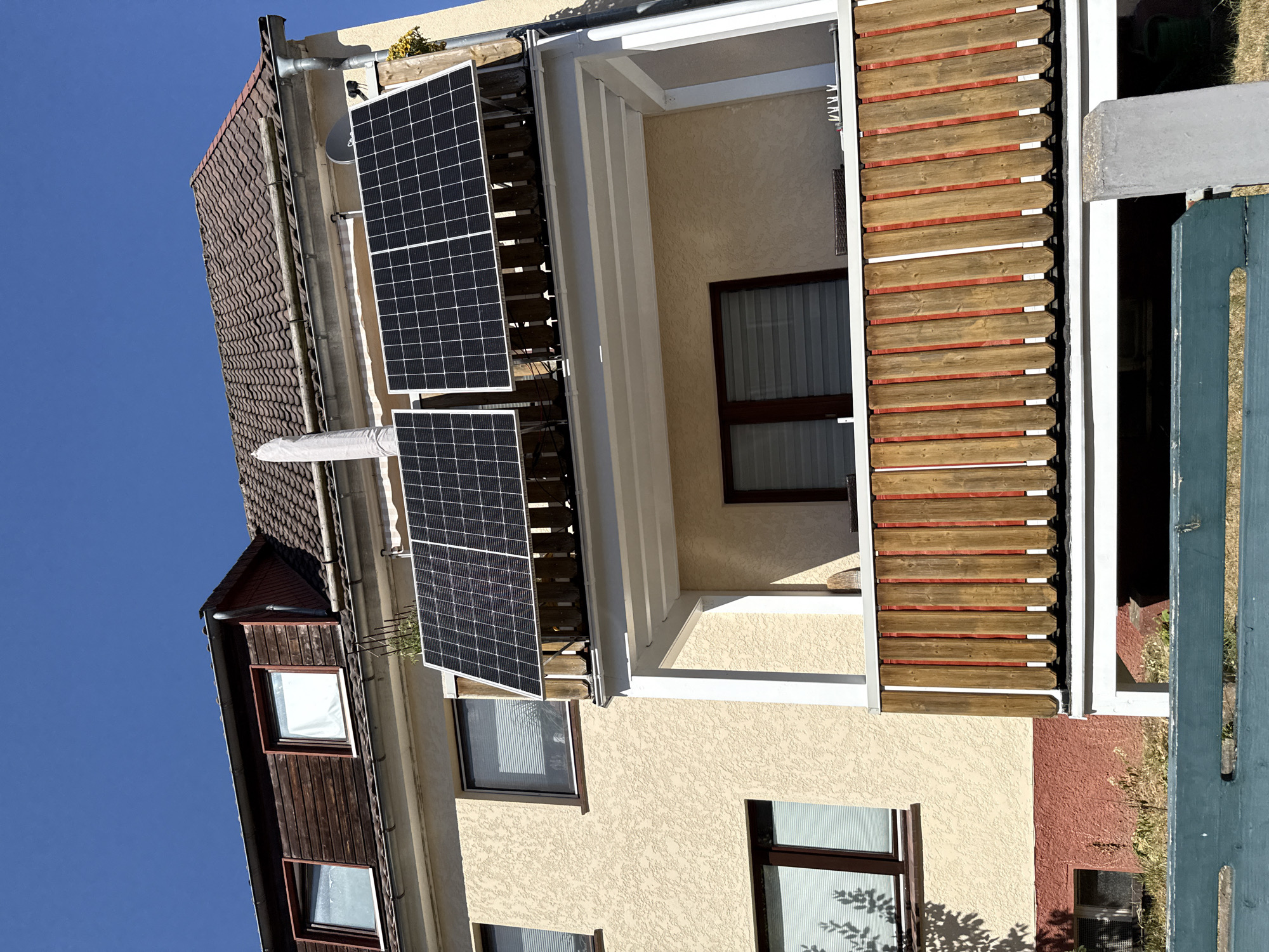 The image shows a residential building in Bad Sachsa, Germany, on a clear, sunny day. The building has a light yellow stucco facade and a brown tiled roof. On the balcony, there are two solar panels mounted on wooden supports. The balcony railing is made of vertical wooden planks with red backing. There are windows on the building, some with white blinds. To the left of the building, there is a section of red-colored lower wall and some dry grass and vegetation. A dark green wooden fence is visible in the foreground.
