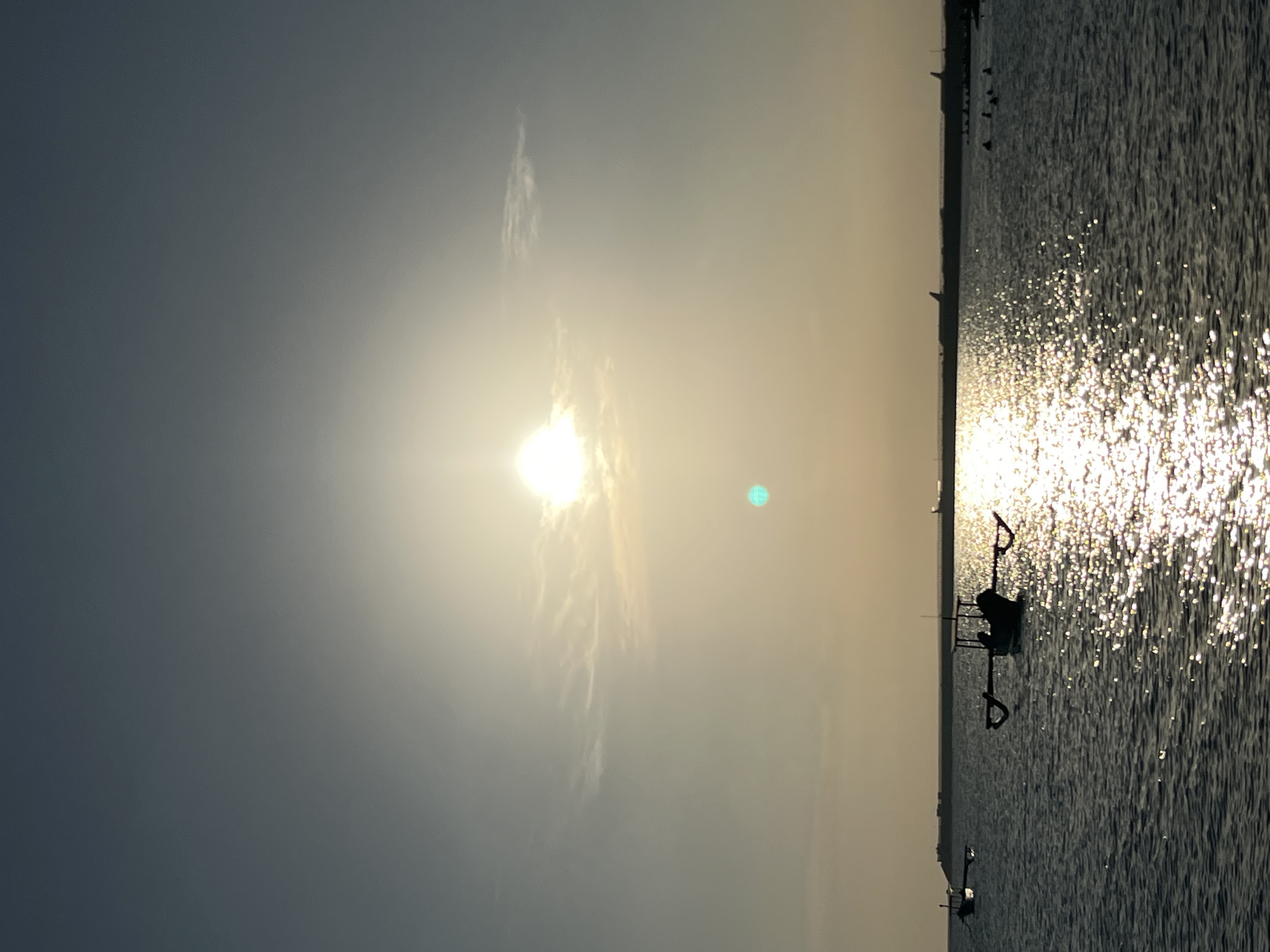 The image depicts a serene sunset scene at Kuta Beach, Bali, Indonesia. The sun, a bright orb, hangs in the hazy sky, casting a shimmering golden reflection on the gentle waves of the ocean. In the foreground, traditional outrigger boats, known as "Jukung," are moored on the water, their silhouettes stark against the luminous sea. A few figures are visible in the distance, seemingly enjoying the evening by the water. The horizon is marked by a long, low structure, possibly an airport runway, with the faint outlines of airplanes suggesting its presence. The overall atmosphere is tranquil and picturesque, characteristic of a Balinese sunset.
