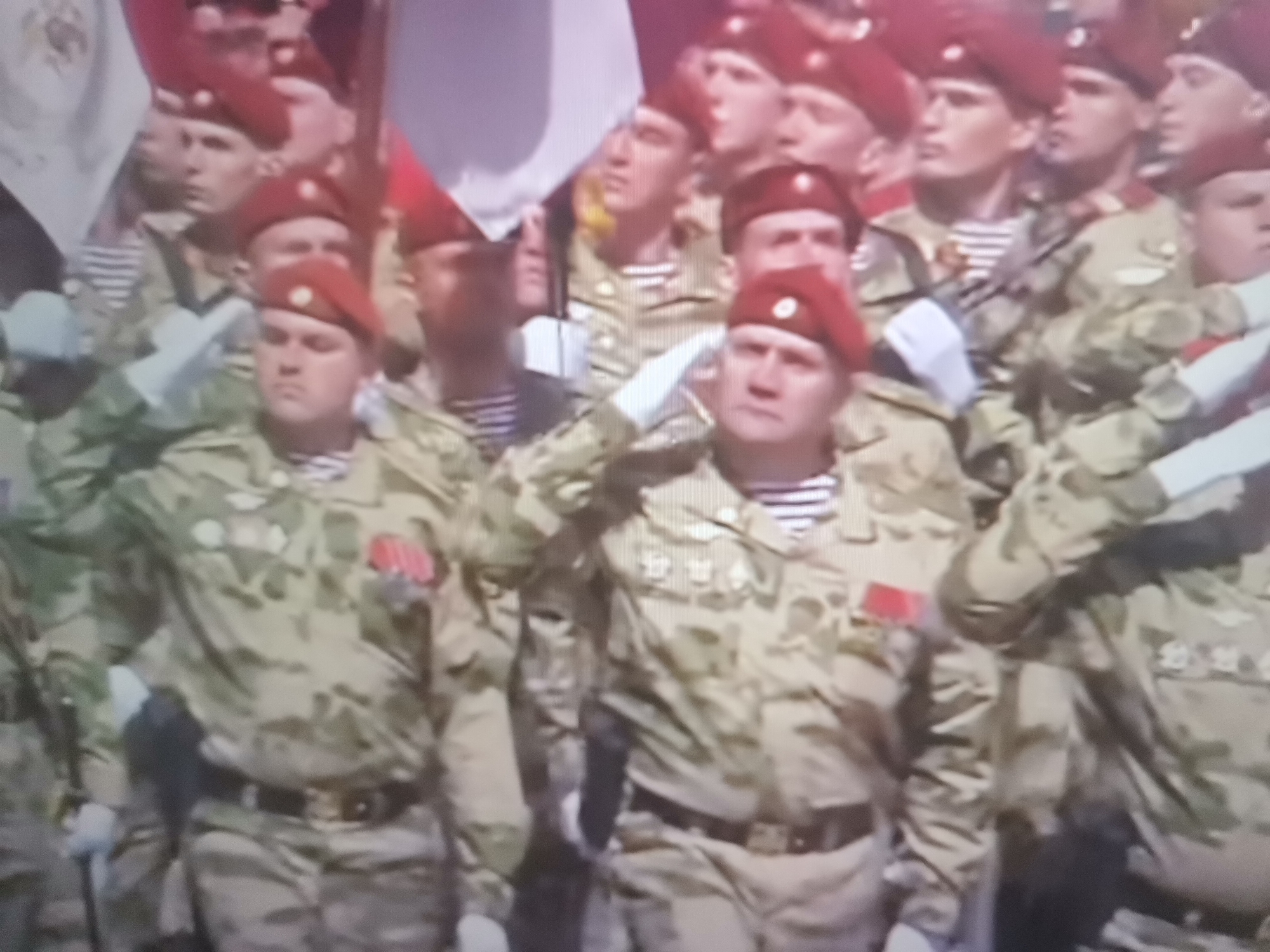 The image depicts a group of soldiers in formation, appearing to be participating in a parade. The soldiers are wearing camouflage uniforms, maroon berets, and striped undershirts. Many of them are saluting. They are armed with what appear to be rifles. The background is blurred but suggests an outdoor setting with flags and possibly buildings. The soldiers' attire and the organized formation indicate a formal military event. The location of Belaazyorsk, Belarus is not visually evident from the provided image.