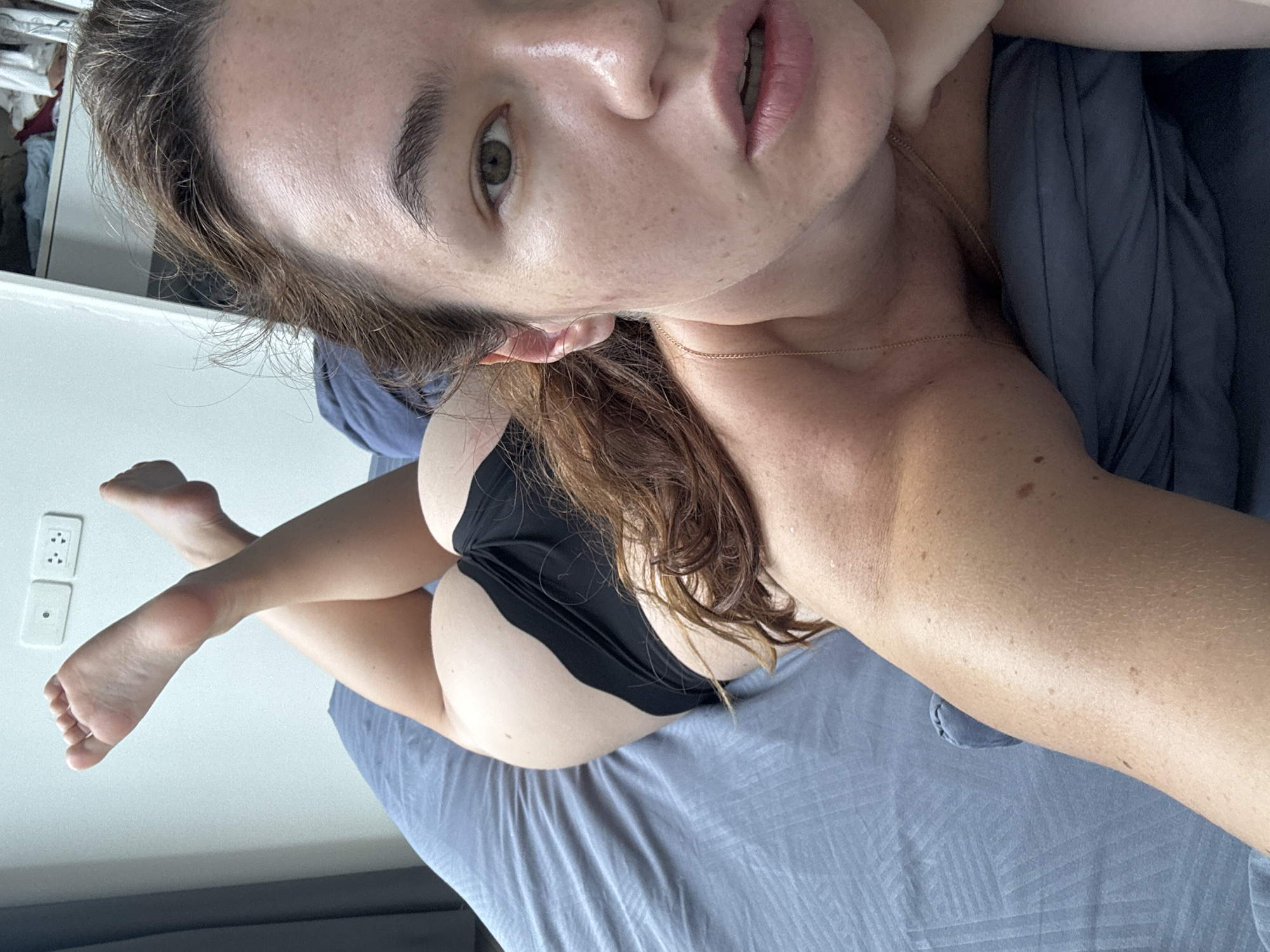A woman is lying on a bed in a bedroom in Ratsada, Thailand. Her body is angled to the side, with her head towards the foreground. She is wearing black underwear. Her legs are crossed at the ankles and bent upwards. The room is dimly lit with natural light coming from a window on the left. There are electrical outlets on the wall behind her. The bed is covered with a blue sheet.