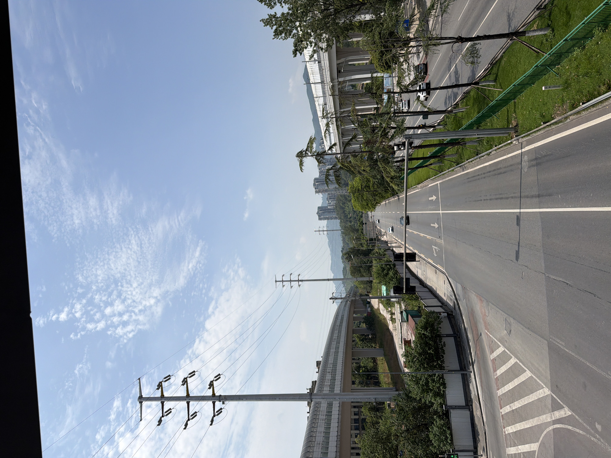 The image depicts a wide, multi-lane asphalt road in an urban environment under a clear blue sky with scattered white clouds. A large elevated structure, likely a transit system or elevated highway, with a translucent roof and white supports, runs along the left side of the road. Tall, grey utility poles with electrical wires are prominent on both sides of the road. Lush green trees line the right side of the road, with some scattered on the left. Several cars are visible on the road, indicating moderate traffic. Buildings, including tall high-rise structures, are visible in the distance behind the elevated roadway and trees. The scene appears to be a typical urban landscape, possibly within a developed district of Tiaodeng, China, given the architectural style and infrastructure. The perspective suggests the photo was taken from an elevated position, perhaps a bridge or overpass.