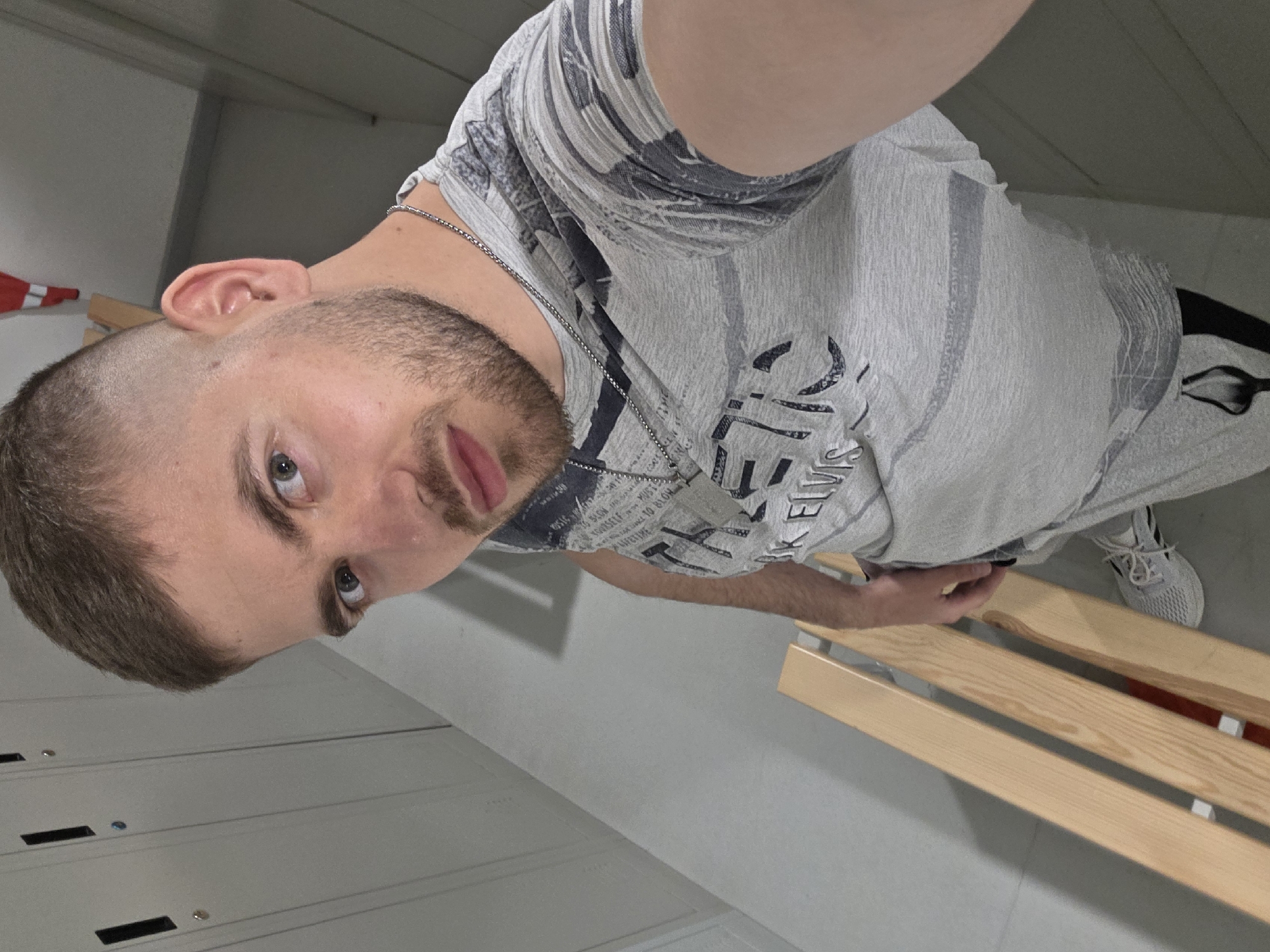 A man is photographed indoors in what appears to be a locker room. He is wearing a gray t-shirt with black and white text, a silver necklace, and gray athletic shoes. He is standing next to a light wood bench. The background includes lockers and a glimpse of a red cone. There is no information in the image to confirm a specific location in Plovdiv, Bulgaria.