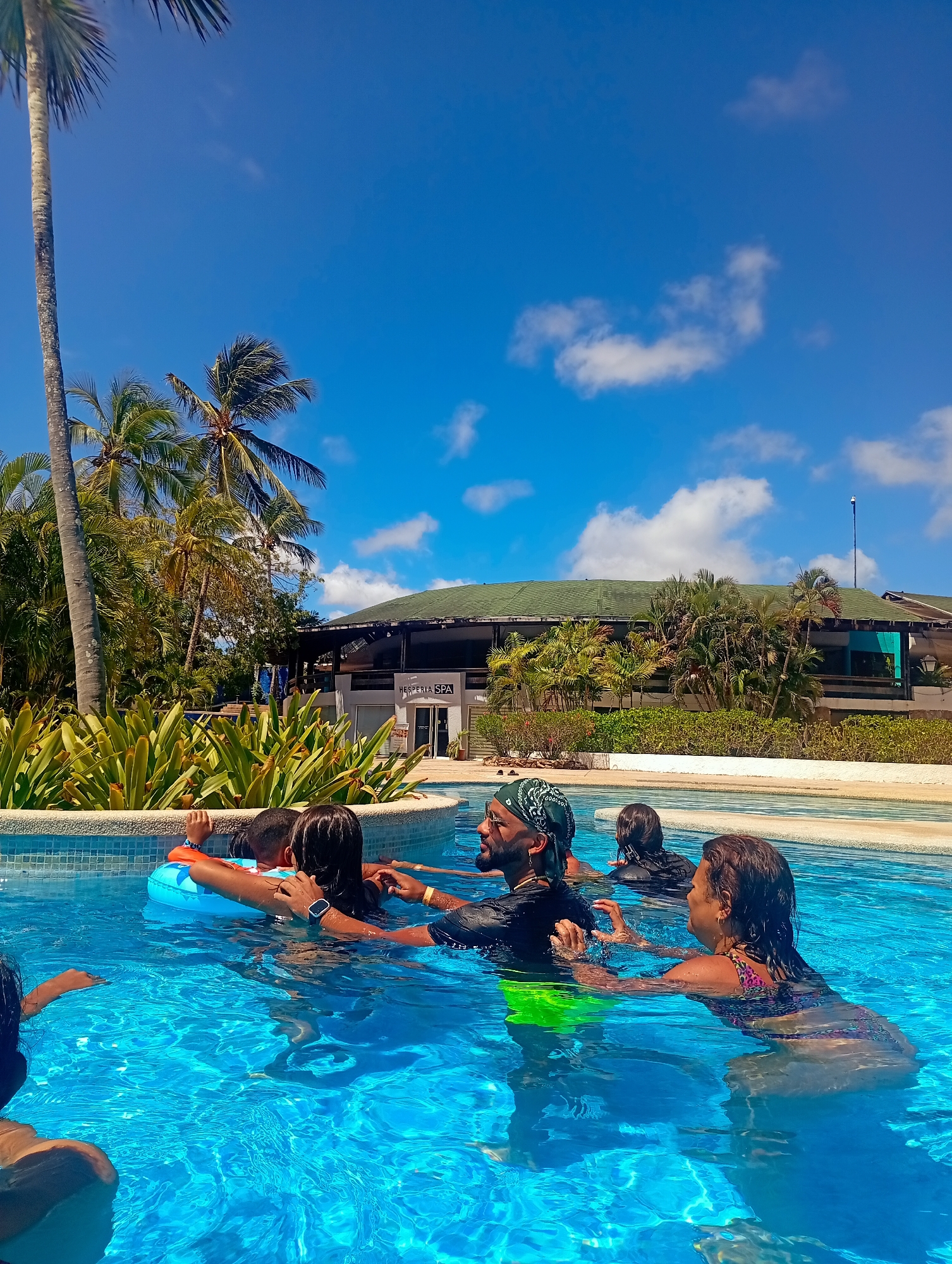The scene is an outdoor swimming pool at the Hesperia Isla Margarita hotel in Playa El Agua, Venezuela, under a bright blue sky with scattered white cumulus clouds. In the background, a multi-story building with a dark green roof and open-air sections displays a sign reading "HESPERIA SPA". A tall pole with an antenna-like structure is visible on the roofline to the right. Lush tropical vegetation, including numerous palm trees and dense green foliage, surrounds the pool and the building.
The clear blue swimming pool occupies the foreground and midground, with a light-tiled edge. Approximately six individuals are partially submerged in the pool. Centrally positioned, a man with a dark bandana, beard, and sunglasses wears a black long-sleeved top and neon green swim trunks, looking left. He interacts with a child to his right (viewer's left) who is wearing an orange and blue inflatable swim ring. An individual with a blue smartwatch on their wrist is adjacent to this child. To the man's left (viewer's right), a woman with dark hair, wearing a patterned swimsuit, is partially submerged and faces left with her hands slightly raised. Additional individuals are partially visible further within the pool, engaged in what appears to be a group activity or instruction.