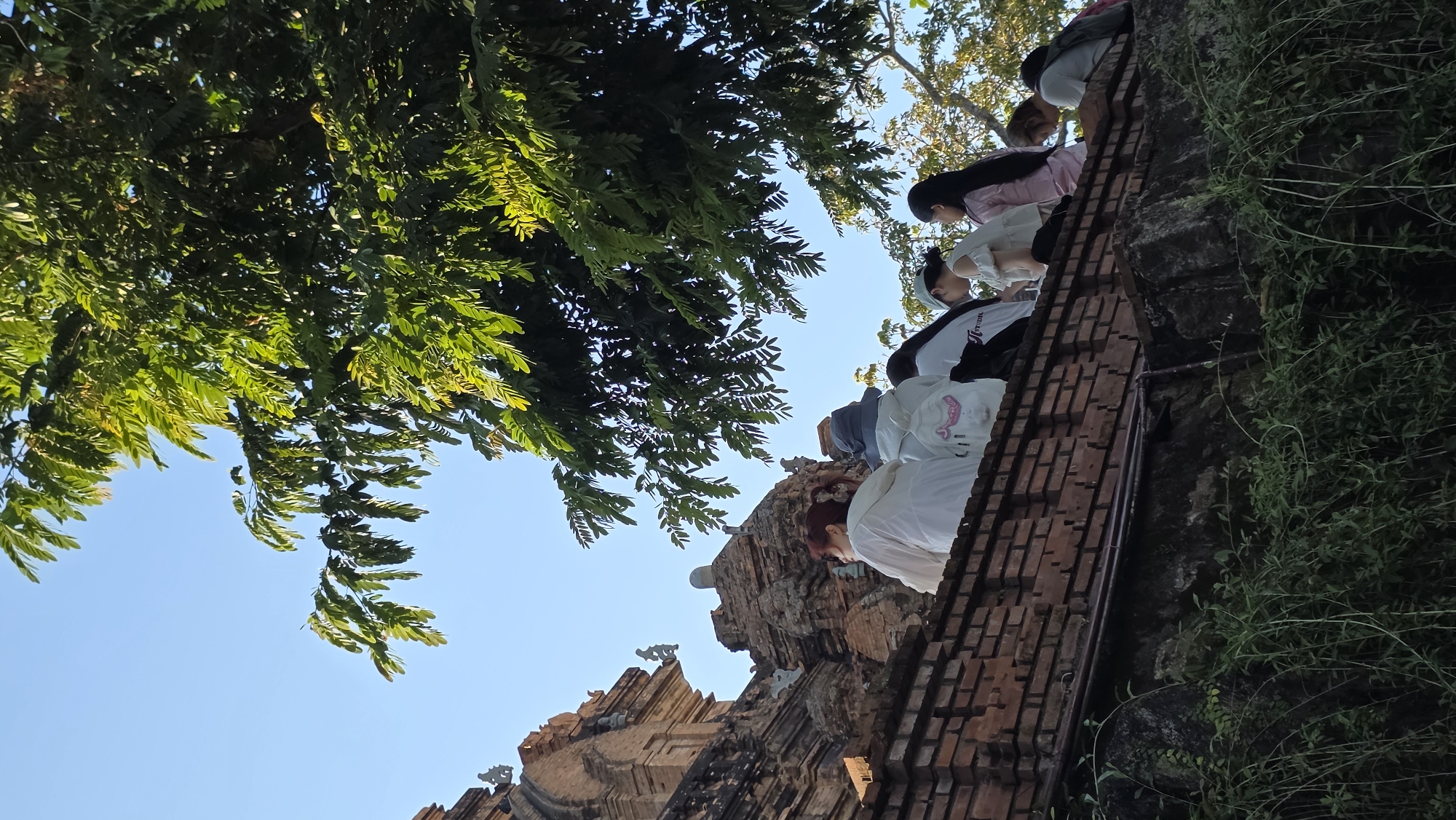 The image shows a low-angle view of Po Nagar Cham Towers, a temple complex in Nha Trang, Vietnam. The foreground features a brick structure with visible brickwork, suggestive of a wall or the base of a tower. At least four people stand on the structure. The background includes tree foliage in the upper left corner, with a patch of clear blue sky. The sky indicates daylight conditions. The camera angle creates a diagonal composition, drawing the eye from the lower left to the upper right.