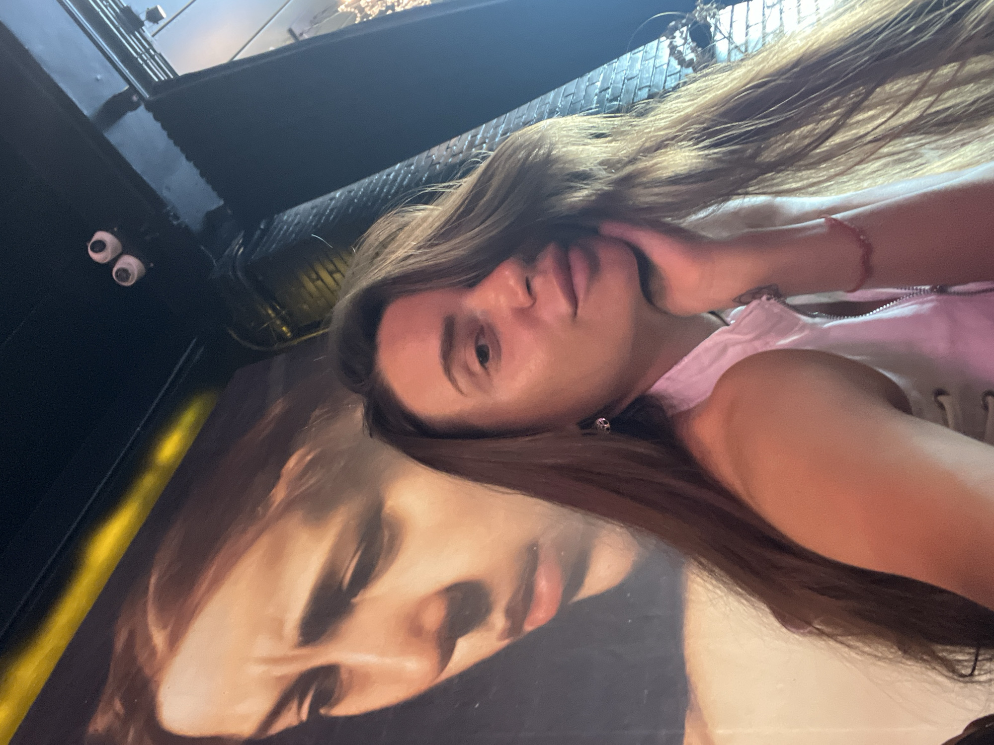 A woman is taking a selfie in a dimly lit indoor setting. She has long brown hair and is wearing a white top. Her right hand is supporting her chin as she looks towards the camera. In the background, a large painting of a woman's face is visible, with a yellow stripe across the top. There are also some security cameras mounted on the ceiling. The overall impression is that of a casual moment captured in a public space. It is not possible to determine the specific location within Ko Samui District, Thailand, based on the visual information provided.