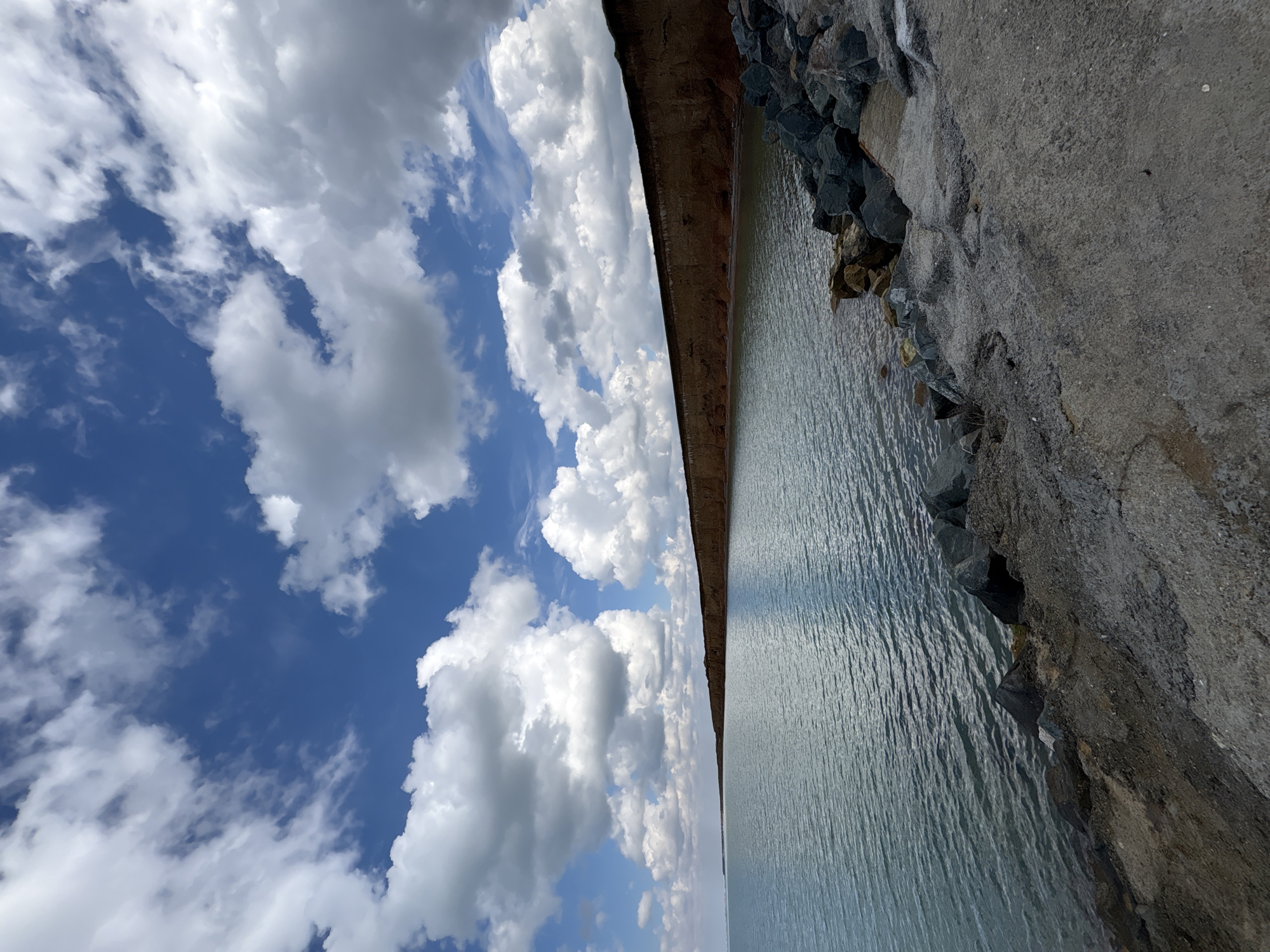 The image shows a body of water next to a concrete structure, possibly a dam or a breakwater, under a blue sky with cumulus clouds. The concrete structure is lined with large rocks along the edge. The water surface is slightly rippled, reflecting the sky. The photograph appears to have been taken at an unusual angle. The location is Николаевское сельское поселение, Ukraine.