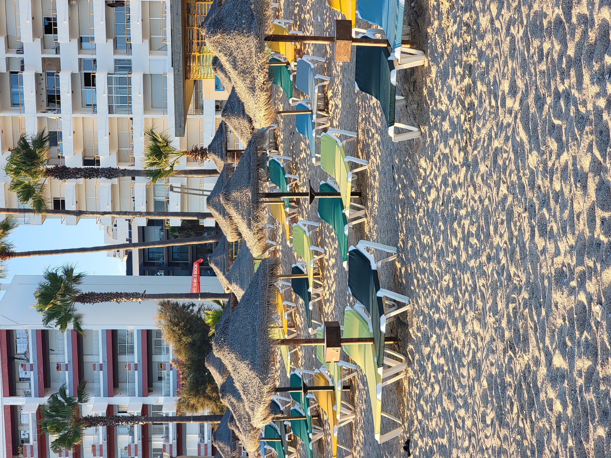 The image captures an empty beach in Quarteira, Portugal, bathed in the warm light of late afternoon or early morning. Rows of unoccupied thatched umbrellas and lounge chairs are neatly arranged on the sandy shore. In the background, multi-story white buildings with balconies and red accents line the coast, interspersed with tall palm trees. The sand is textured with the imprinted patterns of gentle waves and scattered small pebbles. The scene conveys a sense of tranquility and preparedness for visitors, despite the current absence of people.