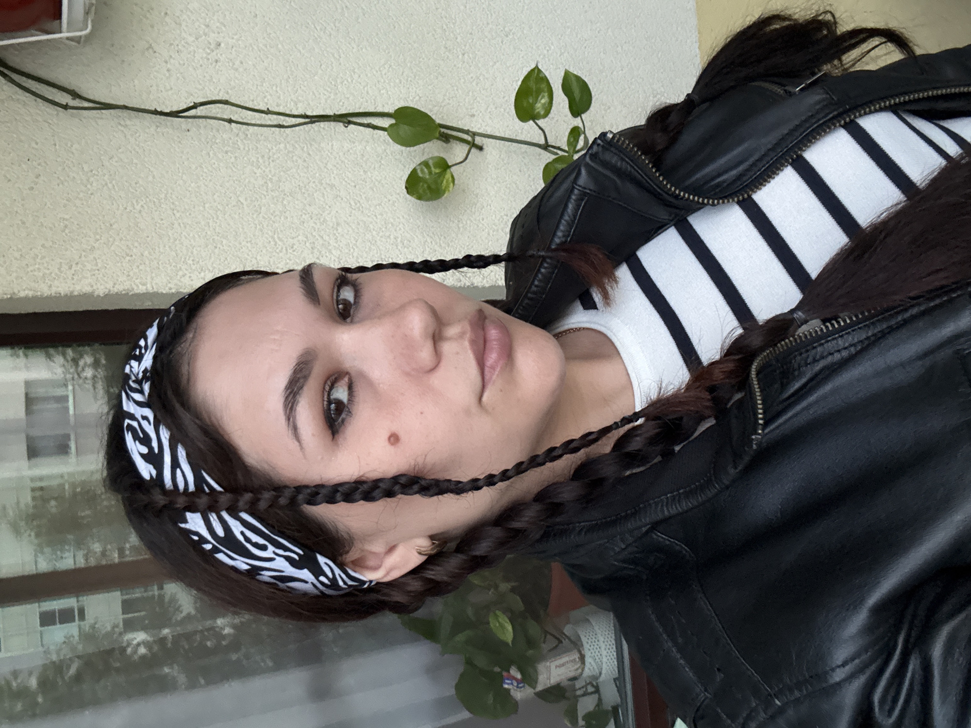 A close-up, vertically oriented, outdoor, medium shot shows a young woman with dark hair in braids. She is looking at the camera with a neutral expression. She is wearing a white and black striped shirt and a black leather jacket. A black and white bandana is wrapped around her head, with the braids coming over her shoulders. A vine with green leaves is visible on the textured, pale yellow wall behind her. The background is blurred but appears to be a building with windows and some plants. The lighting suggests it could be daytime, possibly overcast.