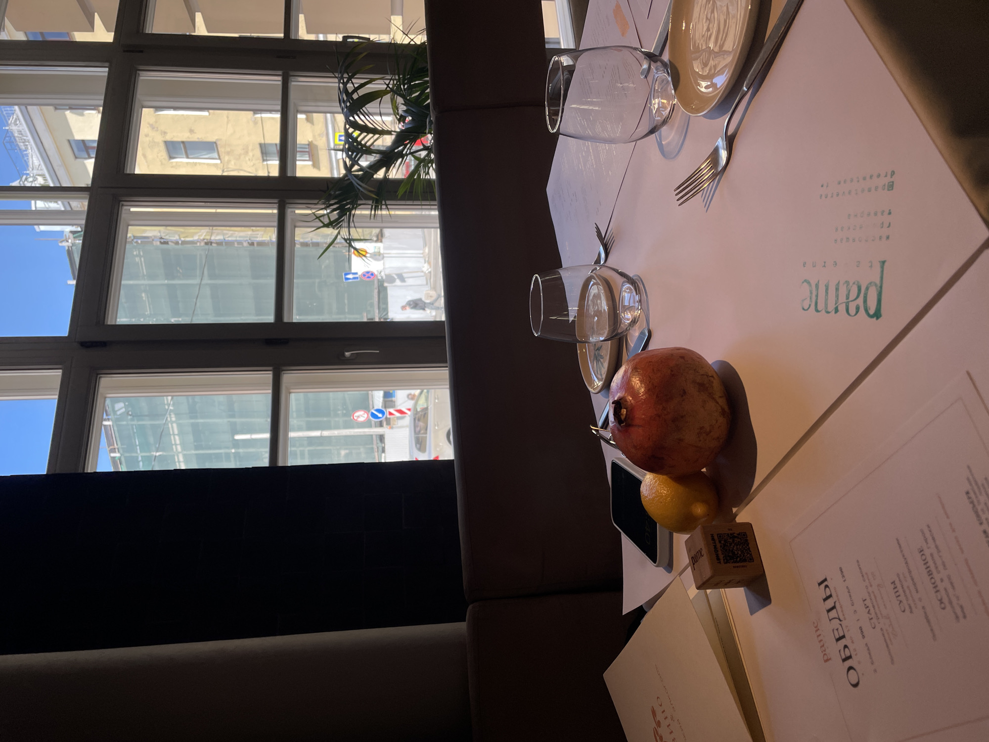 The image shows a table setting at a restaurant, likely in Saint Petersburg, Russia, given the building architecture visible through the window.
The table is covered with a white tablecloth on which a plate, silverware, and two wine glasses are set. A menu card is also on the table with "pame" (Раме) printed on it, likely the restaurant's name. The word "ОБЕДЫ" (Obedi), which translates to "Lunches," and a list of menu items in Russian are printed on it. A QR code is visible on a cube-shaped object.
There is a pomegranate and a lemon on the table as well.
Outside the window, one can see typical Saint Petersburg architecture, with yellow buildings and a clear blue sky. Traffic signs can be seen in the reflection on the glass. The window is multi-paned, and a plant is sitting on the windowsill. A portion of a sofa is visible to the left.