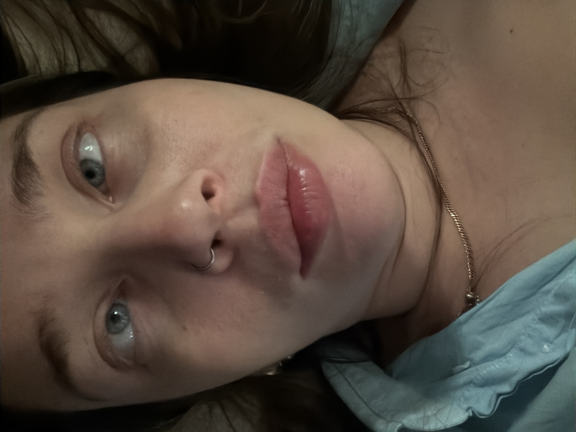 A close-up, angled shot shows a person with fair skin and blue eyes. A silver nose ring is visible in the left nostril. The person's lips appear full and have a slight gloss. A delicate gold chain necklace is visible on the neck. The person is wearing a light blue shirt. The background is dark and out of focus. There is no indication of location in Moscow, Russia.