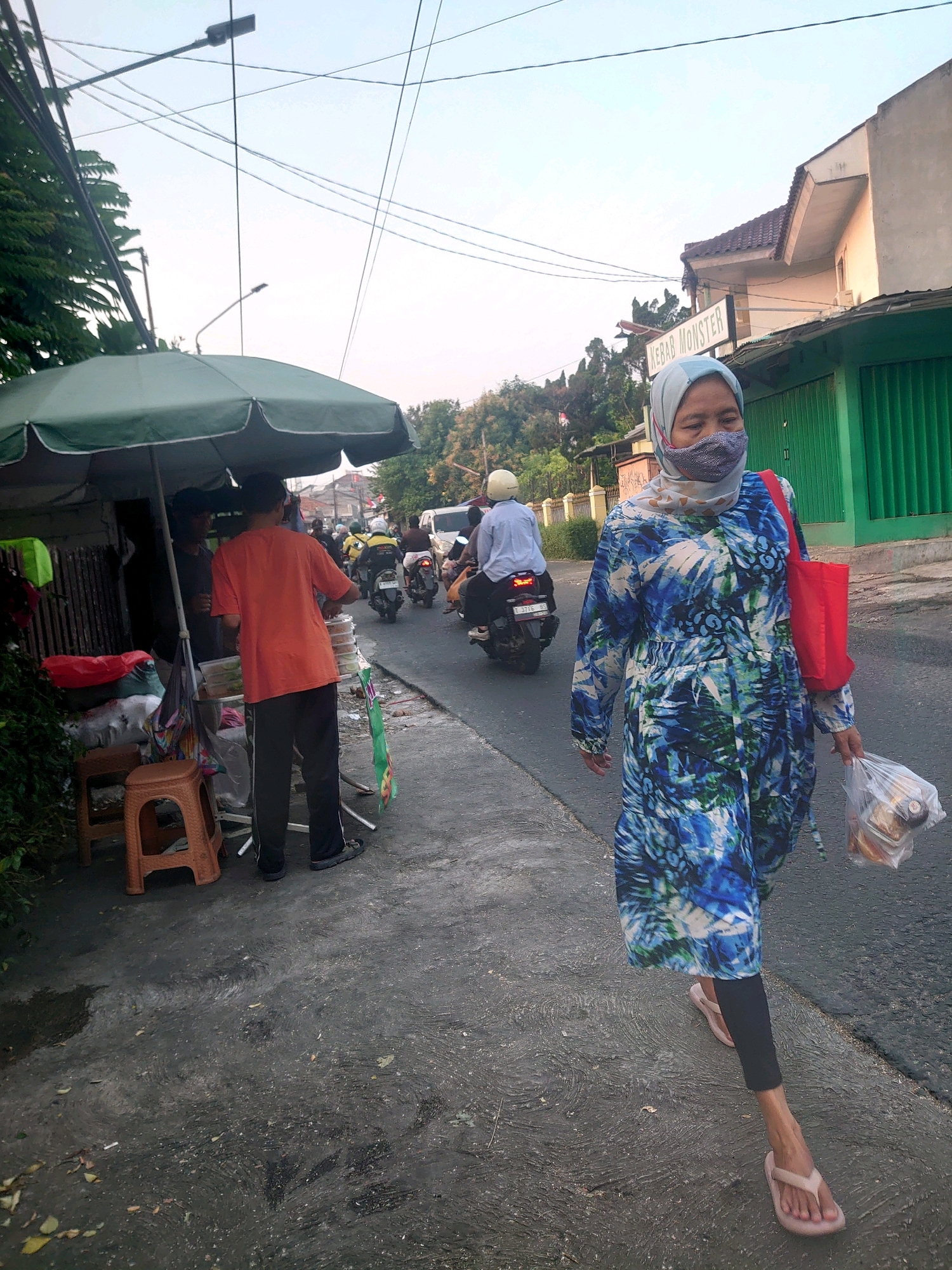 A street scene in Depok, Indonesia, shows a woman walking on the right side of the frame. She wears a blue and green patterned dress, a light blue headscarf, and a patterned face mask. She carries a red bag on her right shoulder and a clear plastic bag containing food in her right hand. In the background, a food stall is visible under a green umbrella, with two men standing behind it. Several motorbikes are also present on the street, with some people riding them. Buildings and trees line the background, and power lines crisscross overhead. The road surface is asphalt with visible water marks, and debris is scattered on the ground.