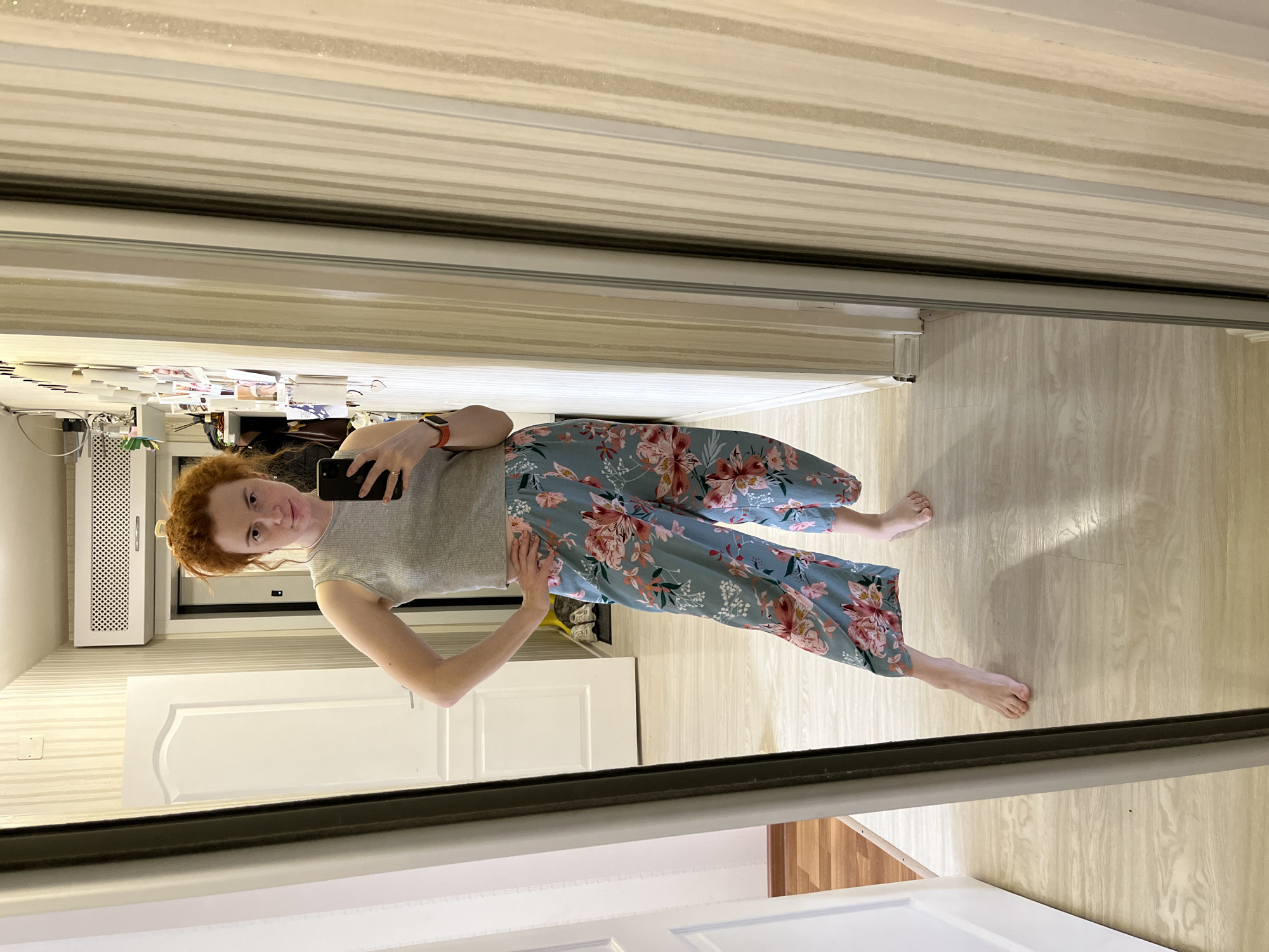 This image features a woman taking a full-body mirror selfie indoors, likely in Chaikovsky, Russia. She has red hair, gathered up, and is wearing a light grey, short-sleeved top and blue wide-leg pants adorned with a vibrant pink and white floral pattern. She holds a black smartphone in her right hand, aimed at the mirror, and her left hand is casually placed on her hip. An orange-banded watch is visible on her left wrist. She looks directly at the camera with a neutral to slightly smiling expression.
The setting appears to be a residential interior, characterized by light-colored wooden flooring and walls featuring horizontal striped wallpaper. The selfie is taken in front of a large, full-length mirror, which seems to be part of a sliding wardrobe or closet door. The reflection shows parts of the room behind the woman: a white paneled door is visible to her left, along with a white wall-mounted heating or ventilation unit with a perforated front above and to the left of the door. A shelf with various items, possibly books or papers, is reflected above her head. The lighting is artificial, suggesting an indoor environment. No explicit text is visible in the image.