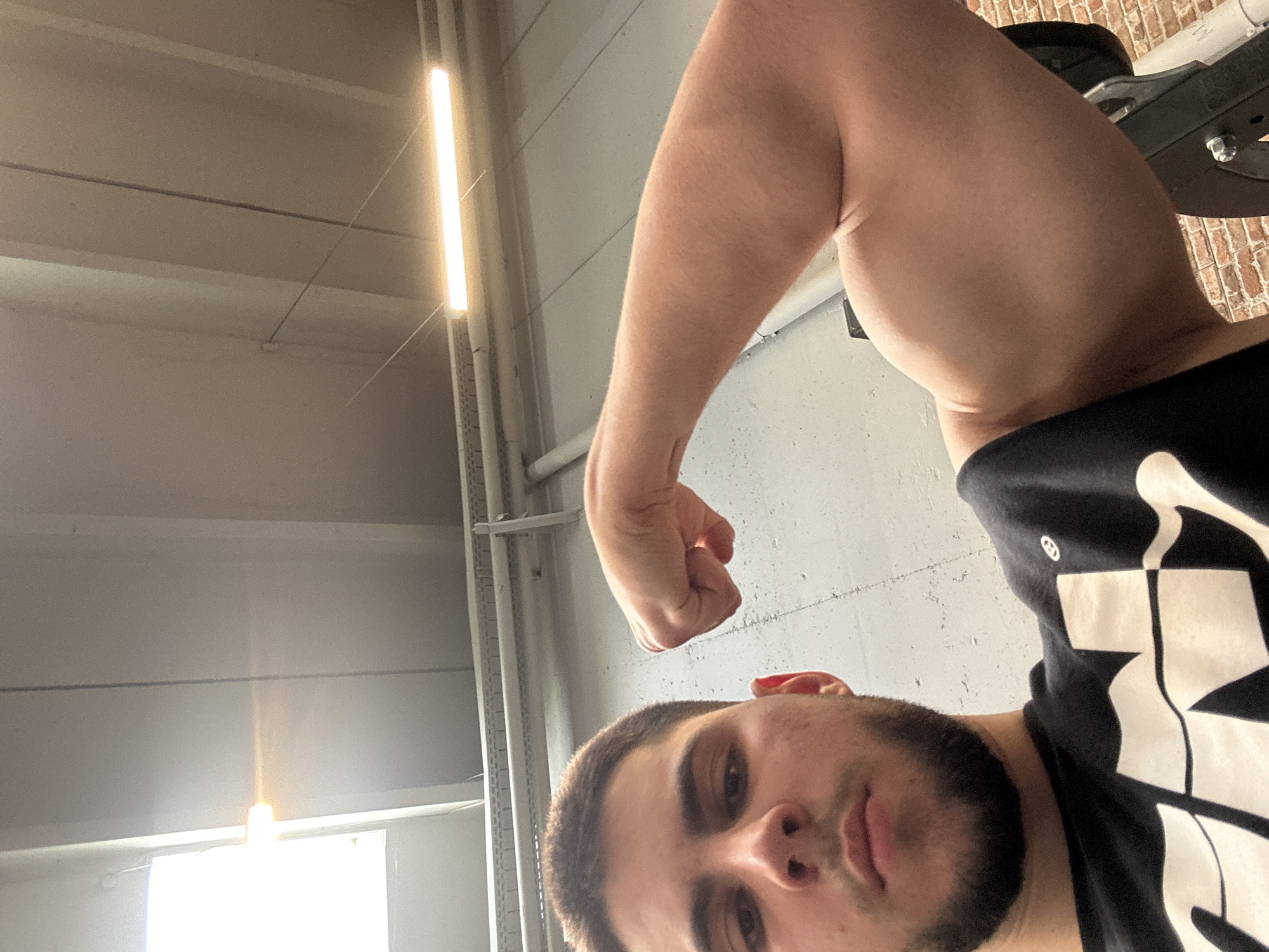 A man with short dark hair and a beard is flexing his right arm, showing his bicep. He is wearing a black t-shirt with a white design. The background is a gym setting with visible ceiling lights and metal pipes. The location is Sofia, Bulgaria.
