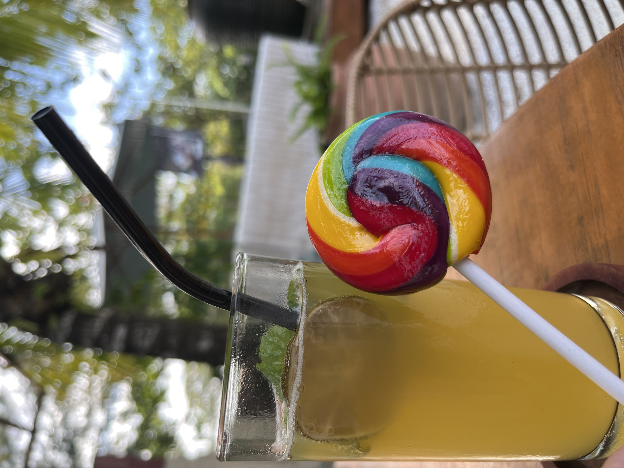 A rainbow swirl lollipop is placed next to a glass of yellow liquid with lime slices and mint leaves. The background is blurred with greenery and hints of structures, suggesting an outdoor setting in a tropical location. The overall impression is a refreshing drink and a sweet treat, possibly at a restaurant or resort.