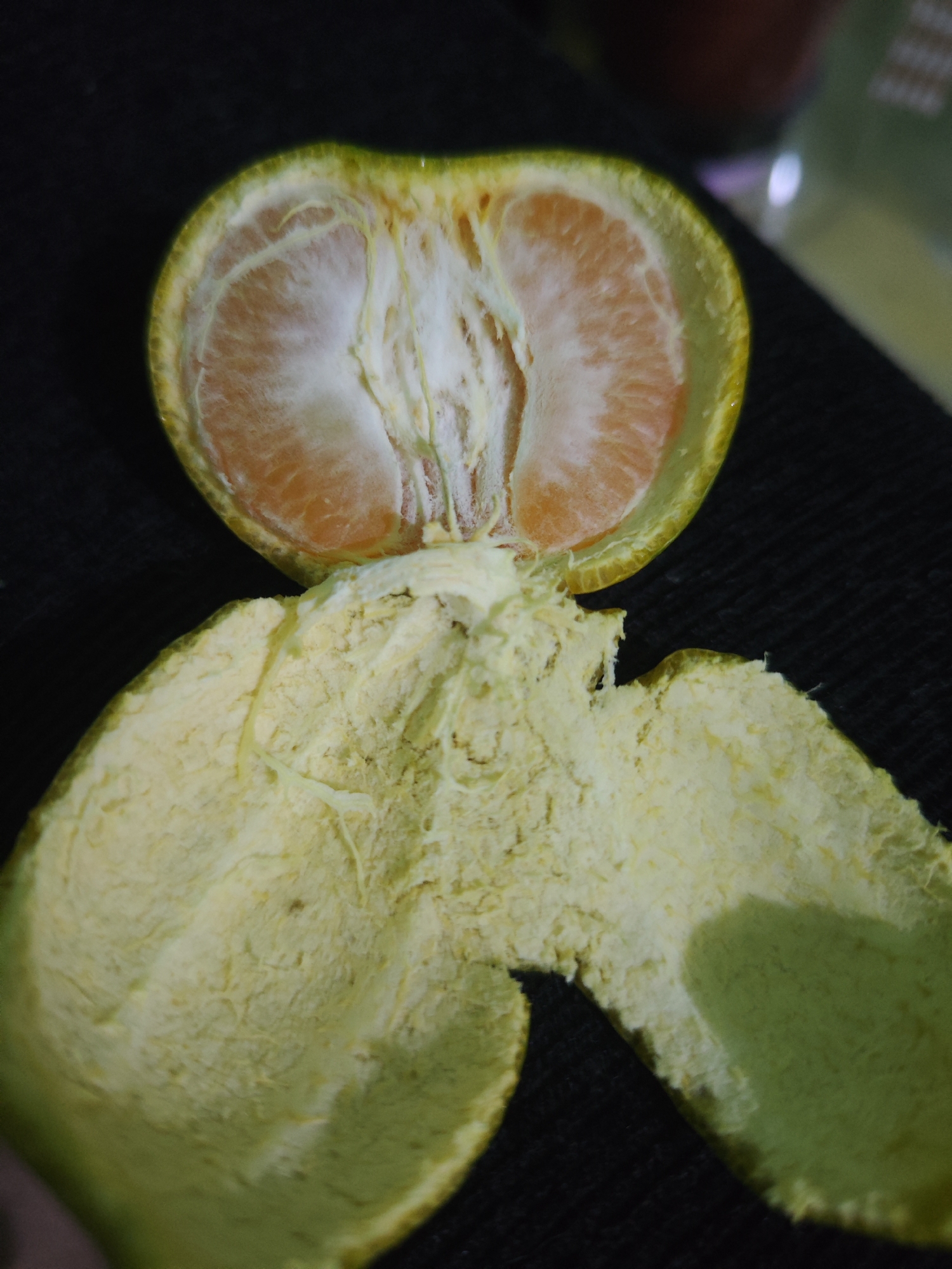 In Sroyo, Indonesia, a peeled orange is displayed against a dark, textured surface. The orange is cut in half, revealing its segmented interior and a pithy white membrane. The peel, separated from the fruit, lies below, showcasing its rough, fibrous texture and pale yellow-green color. The overall lighting is soft, with subtle shadows that add depth to the scene.