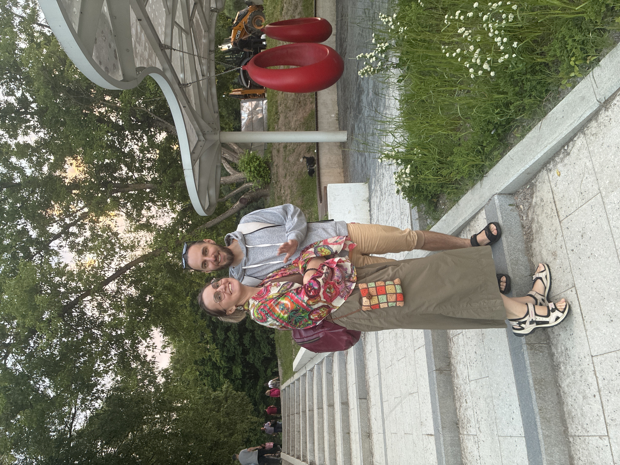 A man and a woman pose smilingly for the camera on a set of wide, light-colored stone steps in an outdoor public space. The image is oriented vertically, with the steps descending from right to left.
The man, on the left, wears a grey hooded sweatshirt, tan shorts, and black sandals. He has sunglasses on his head and a short beard. The woman, on the right, wears a vibrant, multi-colored patterned shirt (featuring red, orange, green, and blue hues), a long olive-green skirt, and white and black sandals. She wears glasses and carries a small, multi-colored crocheted square bag and a larger burgundy shoulder bag. Both subjects are looking directly at the camera with joyful expressions.
Behind the couple, the stone steps continue downwards, with several other blurry figures visible further away. To their right, a patch of green grass with small white flowers lines the path. To their left, a large, modern white or silver architectural structure is prominent, from which two large, red, teardrop-shaped objects are suspended, possibly swings or decorative art installations. Further in the background, a yellow construction vehicle with the text "JCB" visible on its side can be seen. Abundant green trees and foliage frame the upper portion of the image, indicating a park or landscaped area. The lighting suggests it is daytime, likely late afternoon or early evening. The scene conveys a relaxed and happy atmosphere in what appears to be a well-maintained urban park, consistent with a location like Moscow, Russia.