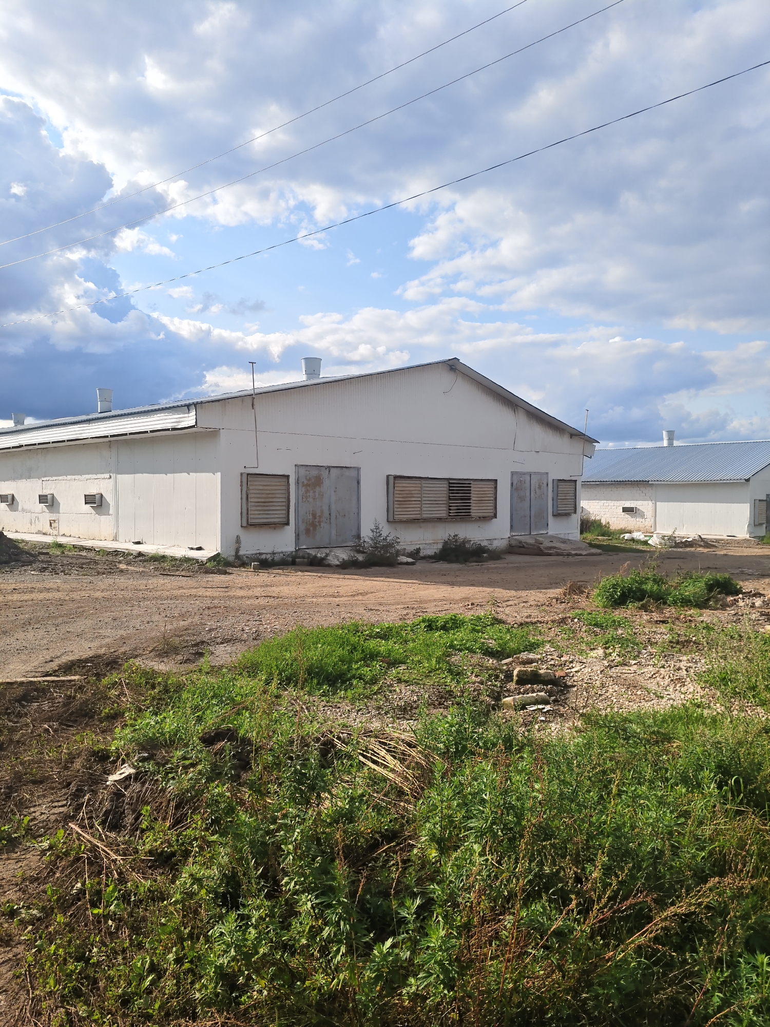 The image depicts an outdoor scene with two white industrial-style buildings under a partly cloudy sky. The building in the foreground is longer and features several windows with slatted shutters and large, weathered grey doors. The building in the background appears similar but is partially obscured. The ground in front of the buildings is a mix of dirt and gravel, with patches of green grass and vegetation growing along the edges and in the foreground. Power lines are visible across the sky. The location is identified as being in the city of Kukuştan, Russia. No people are present in the image.