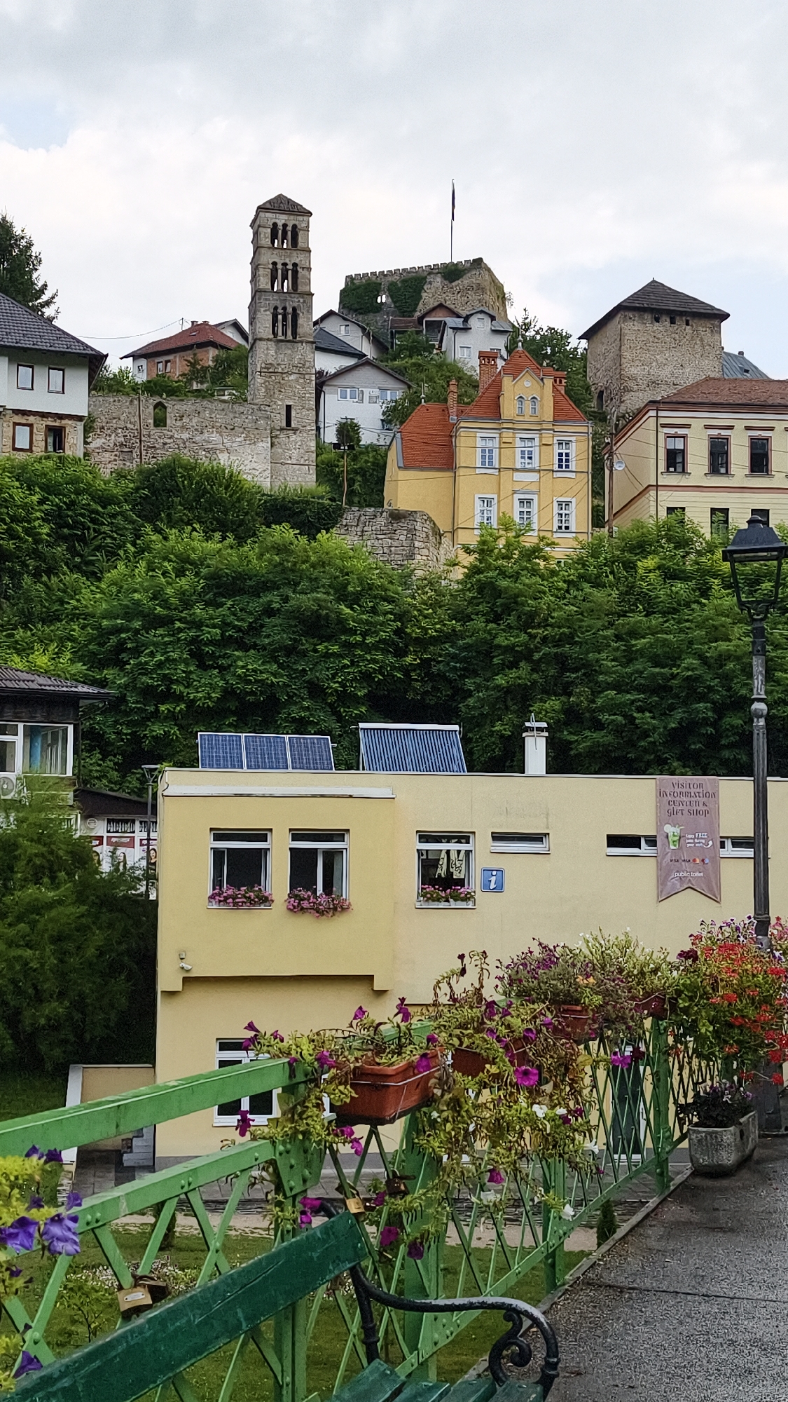 The image depicts a panoramic view of Jajce, Bosnia and Herzegovina, focusing on a hillside dotted with historic architecture and lush greenery. Prominently featured is the St. Luke's Church bell tower, an iconic landmark, situated amidst ancient stone walls that suggest the presence of a fortress or castle. Several residential buildings with red-tiled roofs and yellow facades are scattered across the slope. In the foreground, a contemporary yellow building with solar panels on its roof stands out. A green metal bridge adorned with hanging flower baskets and blooming plants crosses the lower part of the frame, with a park bench visible on it. A lamppost stands to the right, and a banner indicating a "Visitor Information Center & Gift Shop" is affixed to the yellow building. The sky is overcast with scattered clouds.