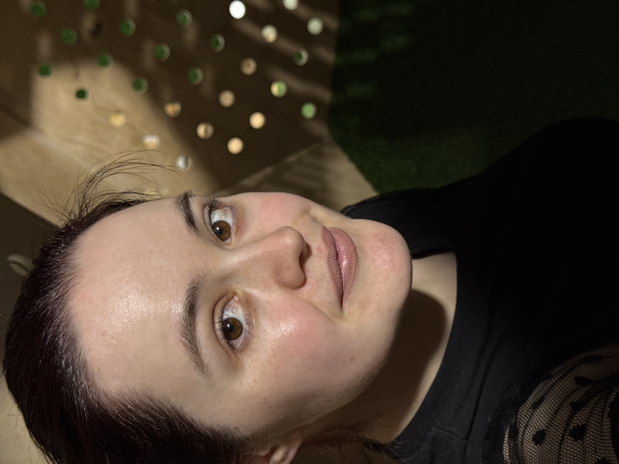 A medium close-up shot captured in Izhevsk, Russia, features a person looking upwards, tilted, and gazing towards the camera. The subject has fair skin, brown eyes, dark eyebrows, and dark hair. They are wearing light pink or nude lipstick. Their expression is neutral to slightly pleasant.
The person is wearing a dark, likely black, top. The visible part of the right sleeve shows a sheer, patterned fabric, possibly lace or mesh, adorned with small polka dots.
The background is dark and out of focus, indicating low light conditions, likely evening or night. Behind the person's head, there's a structure with multiple circular openings or cutouts. Through these openings, various blurred lights (bokeh) are visible, some appearing green and others white or yellowish. Further in the background, dimly lit and blurred dark foliage or grass can be seen. The primary light source appears to be artificial, illuminating the person's face from an elevated position, creating subtle shadows under the chin and along the jawline. There is no visible text in the image.