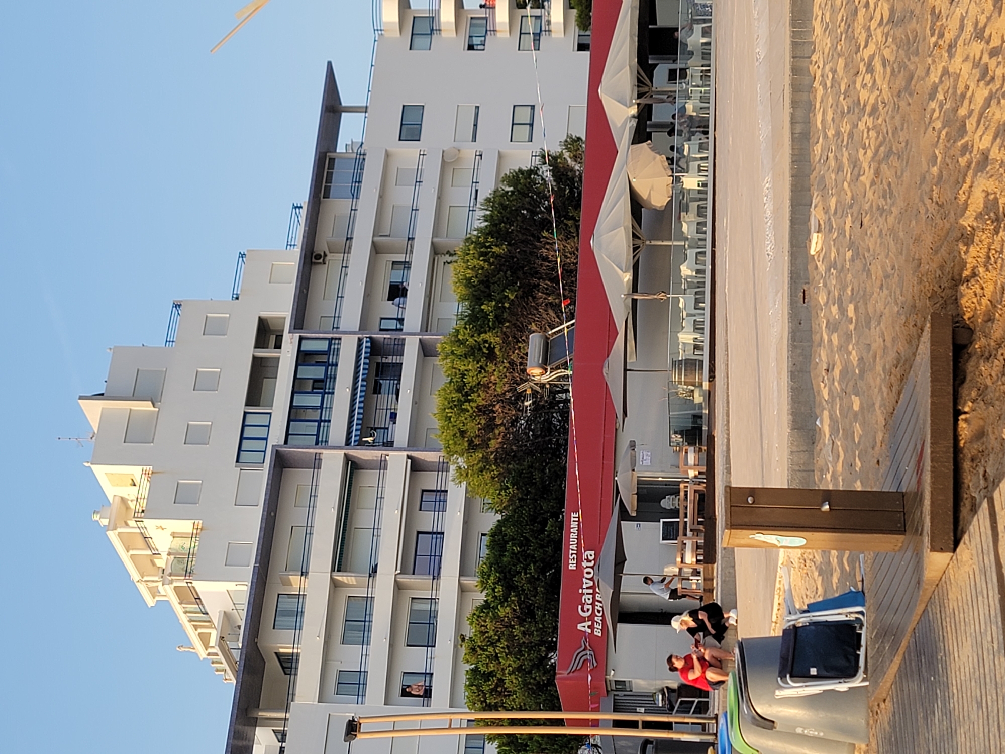 This image captures a sunlit scene in Quarteira, Portugal, likely near the beach. In the foreground, a wooden platform with a shower fixture is set on the sand. To the left of the platform, two blue and grey trash receptacles are visible. A person in a red shirt and another in black are seated on a low concrete wall behind the trash cans, engaged in conversation.
Behind them, a red-awninged restaurant named "A Gaivota BEACH BAR" is situated. Several outdoor tables and chairs are set up under large beige umbrellas in front of the restaurant. A man in a light blue shirt is seen working behind the tables.
Dominating the background is a tall, white, multi-story apartment building with numerous balconies and windows. The building's architecture is modern with stepped levels and blue railings on some balconies. Lush green trees line the area between the restaurant and the apartment building. The sky is clear and bright blue.