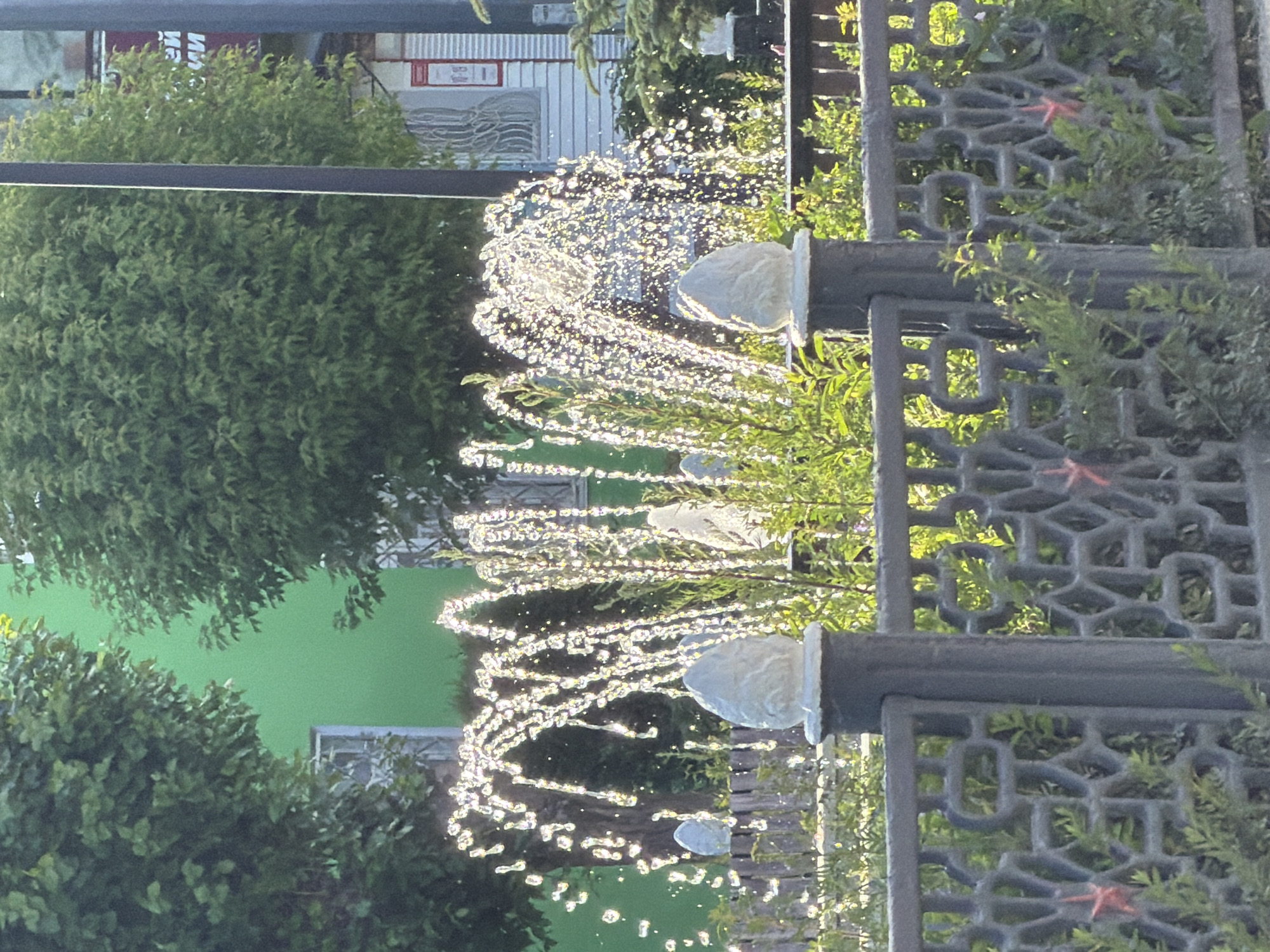 The image depicts a fountain spraying water in arcs, illuminated by sunlight which creates a sparkling effect. The fountain is situated in front of a black metal fence with a decorative pattern and small red star embellishments. Behind the fence and fountain are lush green plants and trees. In the background, a bright green building with white-framed windows is visible, and to the right, a white building with some signage. The overall scene suggests an outdoor public space, possibly a park or a street with landscaping.
