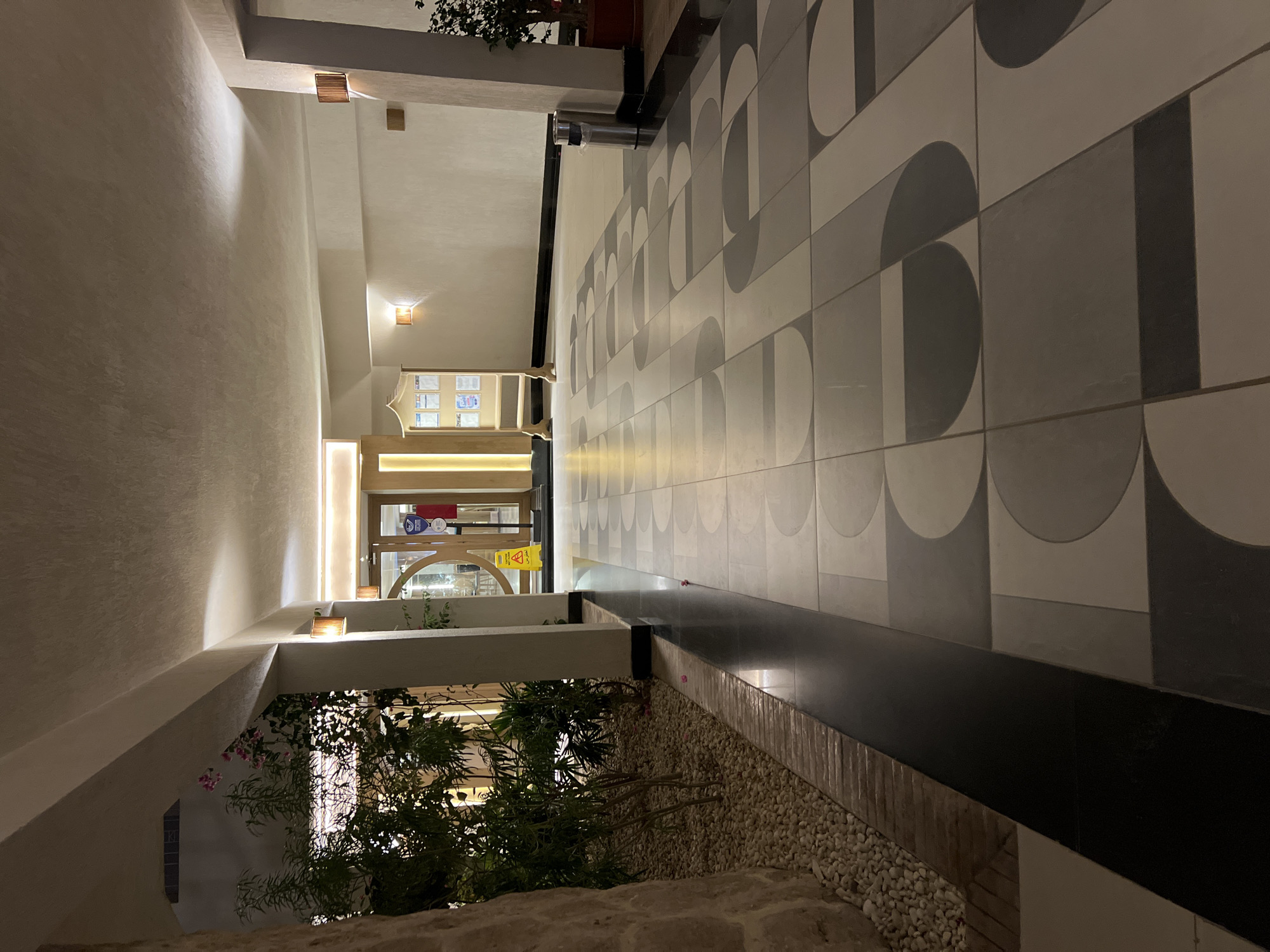 The image depicts an indoor corridor with a patterned tiled floor in neutral tones. On the left, there is a landscaped area with plants and white pebbles. The corridor is illuminated by ceiling lights and wall sconces. Towards the end of the corridor, there are glass doors leading to an outdoor area. A wooden display stand with several papers is visible on the right side of the corridor, along with a silver trash can. The overall scene suggests a hotel or resort hallway in Sharm El Sheikh, Egypt, likely at night given the artificial lighting. There are no people visible in the image.