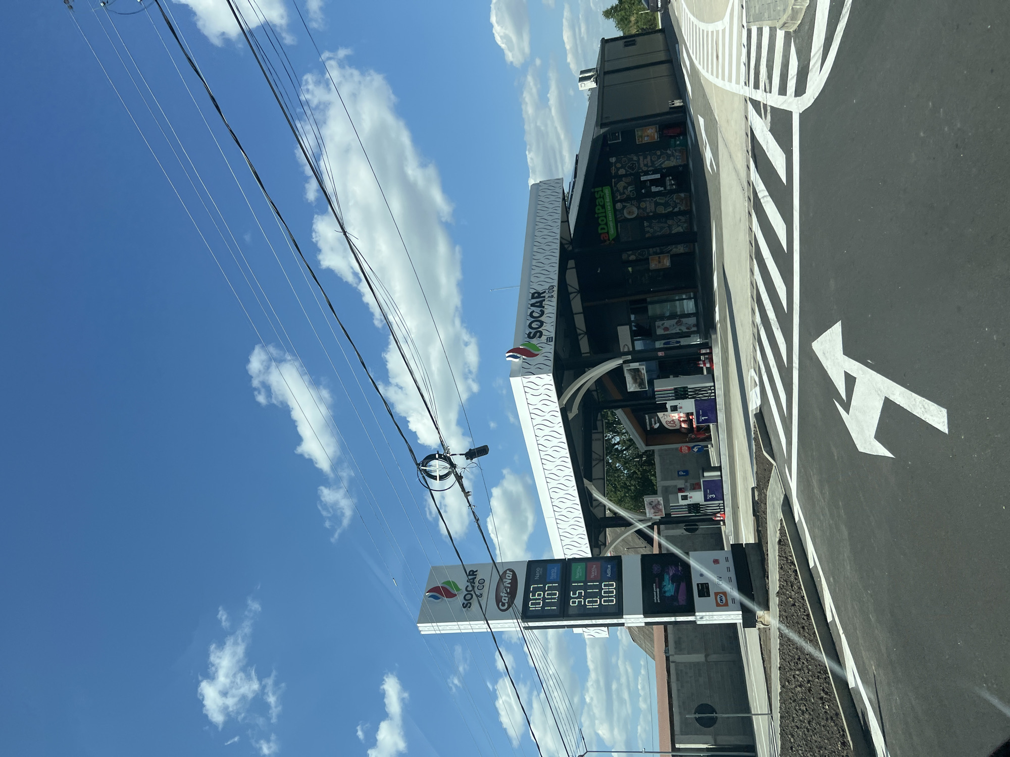 The image shows a view of a SOCAR gas station and convenience store in Sasca Nouă, Romania, on a clear, sunny day with a bright blue sky and scattered white clouds. In the foreground, a newly paved road features white directional arrows and crosswalk markings, indicating a traffic flow.
To the left, a tall, illuminated sign displays fuel prices: 10.67 for Nano, 11.07 for Nano Plus, 9.51 for Diesel, 10.11 for Diesel Plus, and 0.00 for AdBlue. Above the prices, the SOCAR & CO and "Cafe Nar" logos are visible. Further left, a brick building with a brown roof is partially visible.
The central focus is the gas station itself, with a modern, white-tiled roof structure covering the fuel pumps. The building behind the pumps houses a convenience store, with signage including "la Doipasi" and various food-related graphics. Several people are visible in and around the store entrance, appearing to be customers or employees. Fuel pumps are lined up, some with visible pricing displays.
Power lines crisscross the sky above the gas station, with some equipment attached to them. The overall scene depicts a typical roadside service area in a semi-urban or rural Romanian setting.