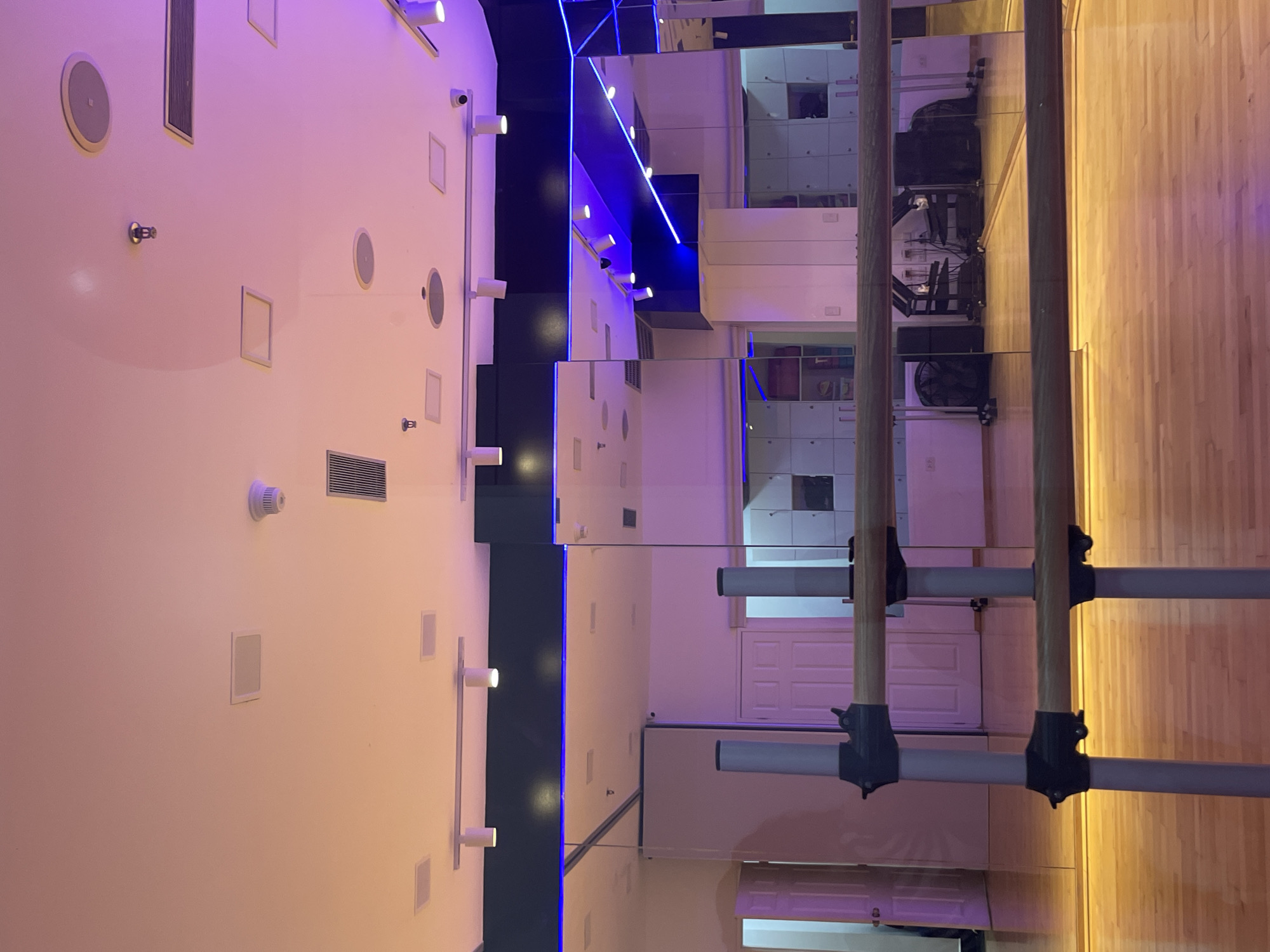 This image shows the interior of what appears to be a dance studio or gym. The studio features mirrored walls, a barre for ballet or exercise, and a wooden floor. One wall is painted white and has various fixtures such as speakers, lights, and other wall-mounted items. The studio is lit with a combination of overhead lights and blue accent lighting. The scene is in Porlamar, Venezuela.