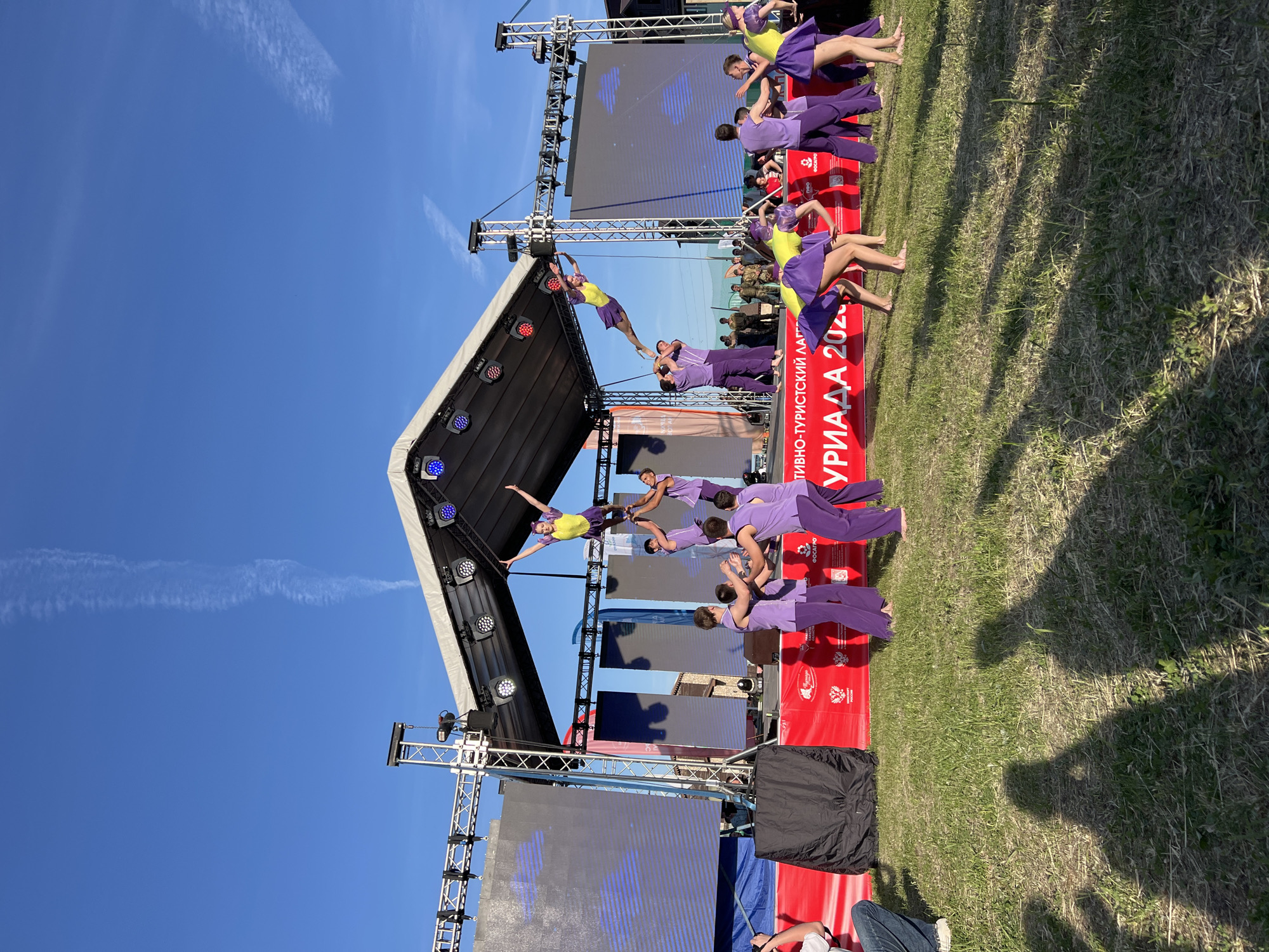 A photo captures a performance scene outdoors on a sunny day in Khvalynsk, Russia. A group of performers dressed in matching purple and yellow outfits is engaging in an acrobatic act on a stage. The stage is set against a backdrop of large video screens, and the event appears to be taking place during the "TIVNO-TOURISTICAL LATTICE 2023", as indicated by the Cyrillic writing on a red banner. The group on the stage consists of approximately 10-15 performers. The scene is set on a grassy area with some spectators present, one wearing blue jeans and white sneakers. The sky is clear blue with a few wispy clouds.