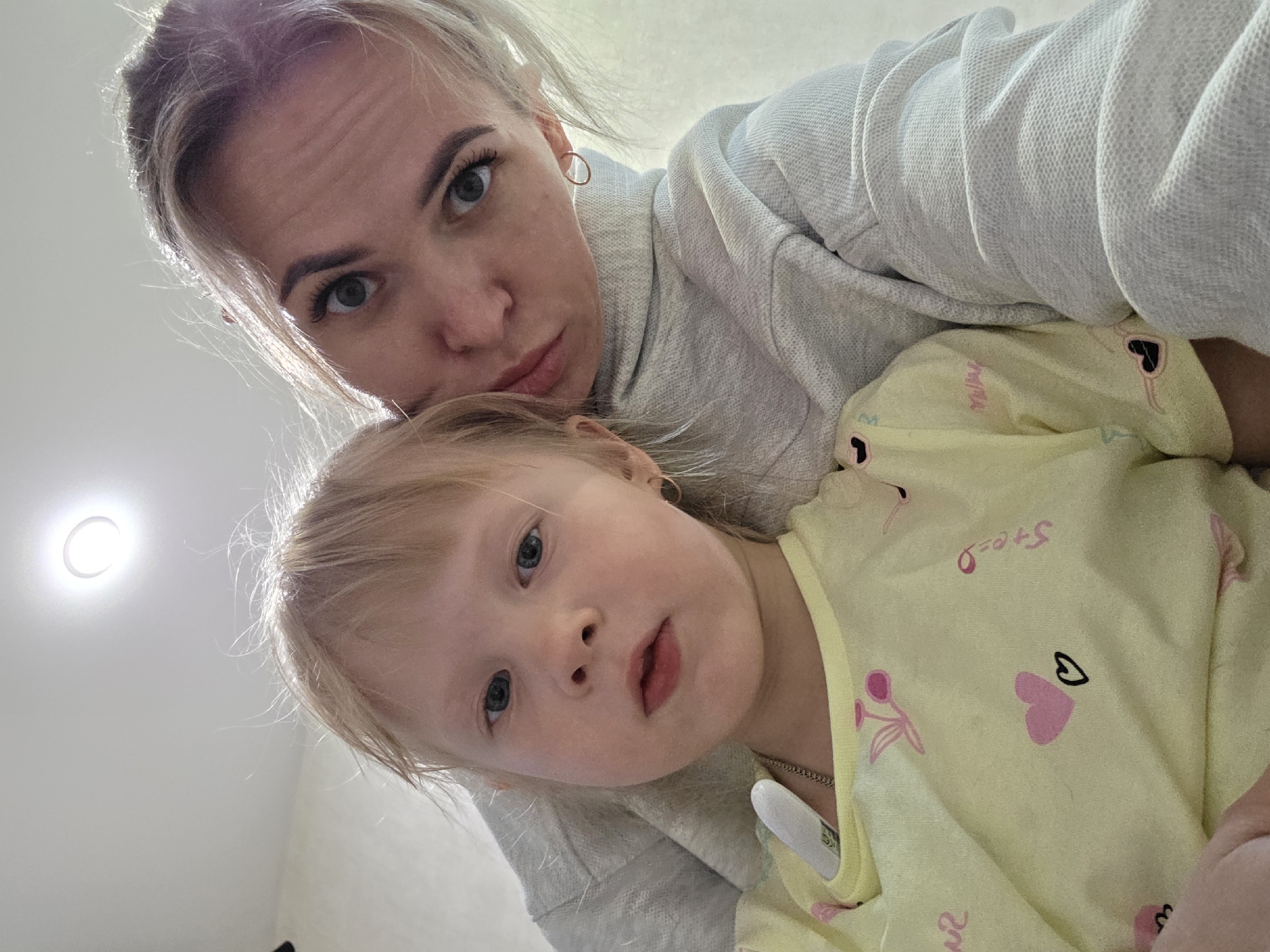 A selfie captures a woman with blonde hair pulled back, wearing a light grey sweatshirt, posing with a young child in a light yellow patterned shirt. The child has blonde hair and is positioned below the woman in the frame. The light source appears to be above, illuminating the faces of both subjects. There is a ceiling light visible in the background. The image was taken indoors, with a plain white ceiling in the background. The location is specified as the city of Chaikovsky, Russia.