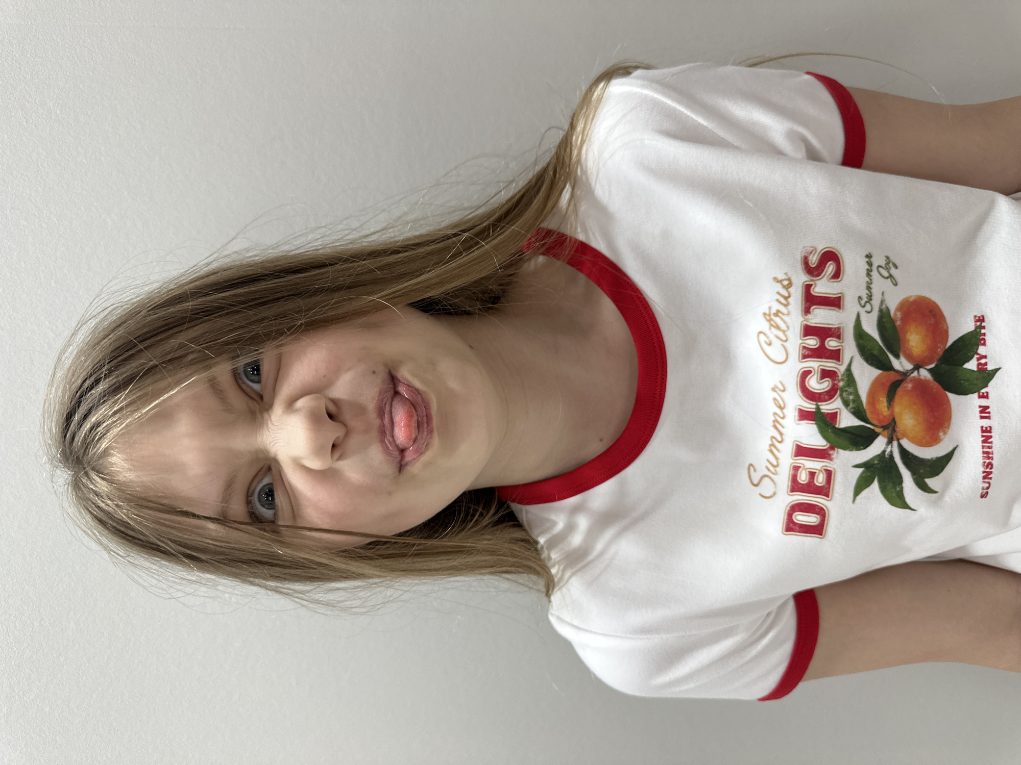 A young girl with light brown hair pulled back from her face, with bangs and some wisps framing her face, makes a funny face with her tongue sticking out and her eyes wide open, scrunching up her nose. She is wearing a white t-shirt with red trim around the collar and sleeves. The t-shirt has a graphic print that includes the words "Summer Citrus DELIGHTS" and an illustration of oranges on a branch with green leaves. Below the oranges, the words "Summer Joy" are printed, and at the bottom of the graphic are the words "SUNSHINE IN EVERY BITE." The background is a plain, light-colored wall, appearing to be a studio or indoor setting, with no indication of the city of Sochi, Russia.