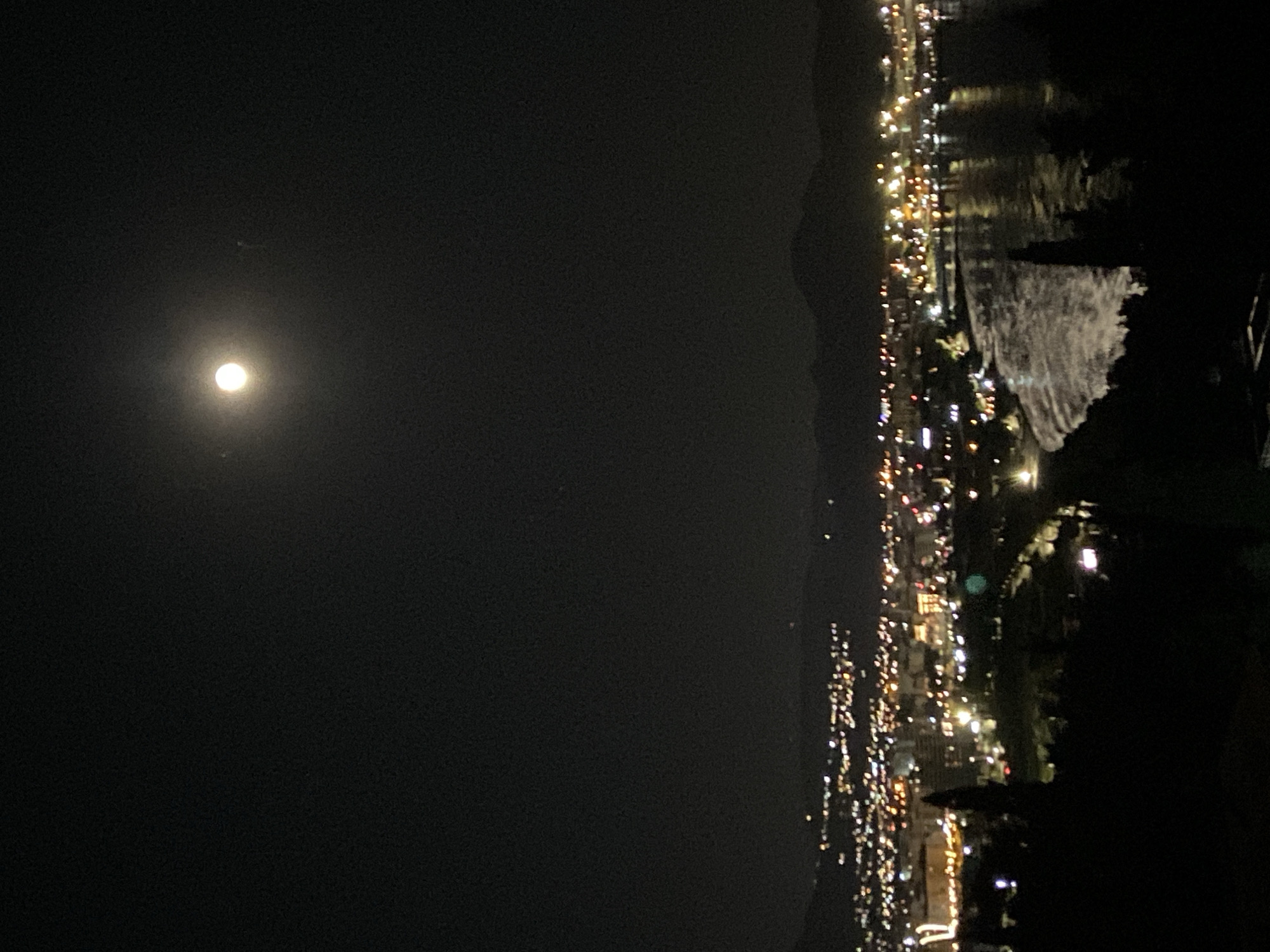 The image depicts a night view of Šušanj, Montenegro, with a bright full moon dominating the sky. To the right of the moon, there is a mountainous landscape visible along the coast. On the right side of the image, a coastal city can be seen, with numerous lights indicating urban development. The lights appear to be reflected on a body of water, possibly the Adriatic Sea. The foreground is dark, with what appears to be vegetation obscuring some of the view. The city lights extend down the coastline, illuminating a dense urban area.