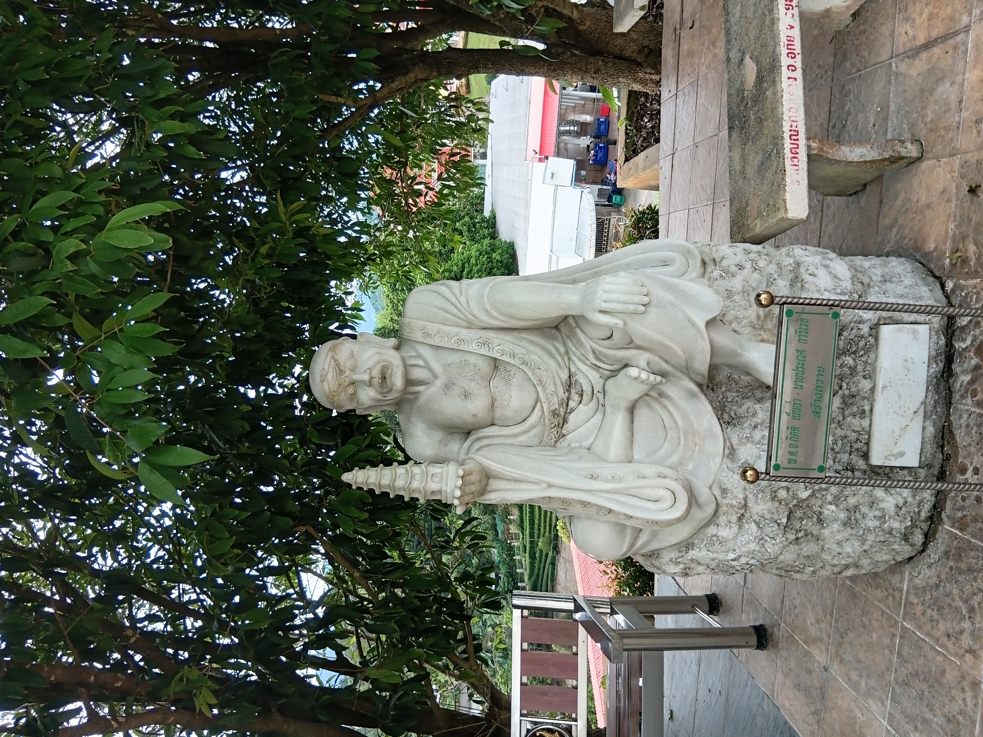 The image shows a seated stone statue of an elderly man holding a small pagoda. The statue is on a stone pedestal, and in front of it is a green plaque with Thai writing and a golden finial on a metal post. The statue is positioned outdoors under a canopy of green trees, with a bench to the left and another stone bench to the right. In the background, there are buildings with red and white roofs, and a terraced landscape, possibly tea or agricultural fields. The scene appears to be in a temple or a park-like setting. The city name "บ้านเหล่าพัฒนา" (Ban Lao Phatthana) suggests a location in Thailand.