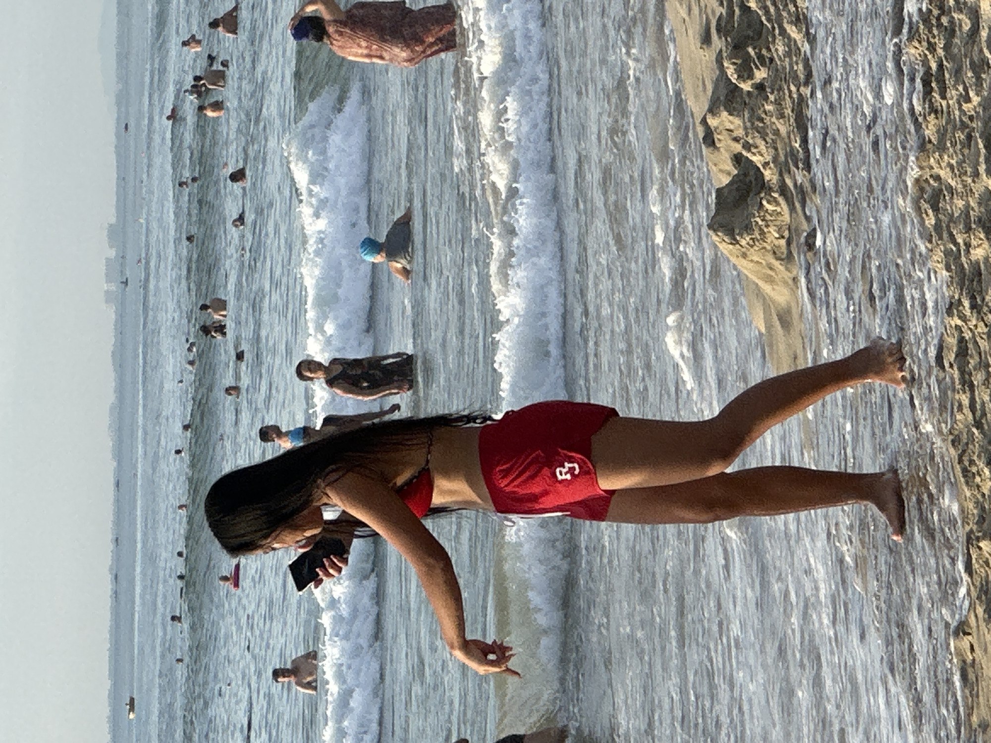 The image shows a crowded beach scene with many people in the water. In the foreground, a young woman with long dark hair is standing in shallow water, wearing a red bikini top and red shorts. She is holding a smartphone in her left hand and her right hand is extended, with her fingers pointing downwards. The water is breaking with small waves around her legs. In the background, numerous individuals are swimming and wading in the ocean. Further in the distance, the ocean stretches to the horizon, with some buildings and possibly a hazy cityscape visible on the shoreline. The sky is a pale, overcast color. The overall impression is a busy summer day at the beach, likely in a coastal city like Đà Nẵng, Vietnam, given the presence of many people enjoying the ocean.