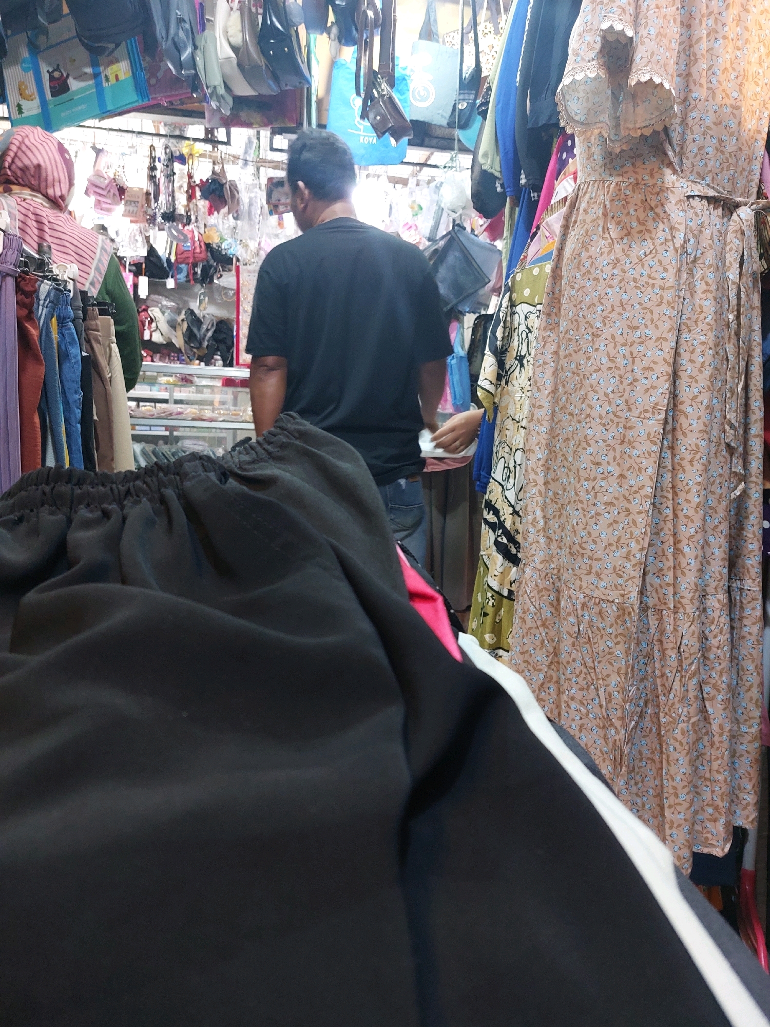 In a market in Sumuragung, Indonesia, a man wearing a black t-shirt stands facing away from the camera, looking at clothing racks. In the foreground, a pair of black pants with white stripes are visible, partially obscuring the lower half of the scene. To the right of the man, a woman's hand is visible, interacting with a receipt or paper, possibly at a checkout counter. The market is densely packed with various clothing items, including dresses, bags, and other garments, displayed on racks and hung from the ceiling. The overall environment suggests a bustling and crowded marketplace.